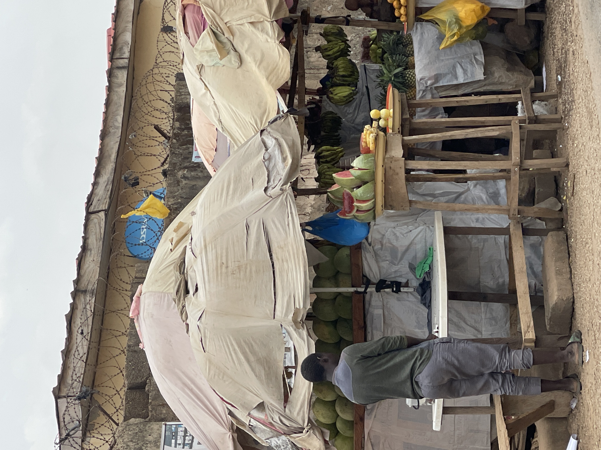 This media file depicts an outdoor fruit stall, likely situated on a street or alleyway in Abuja, Nigeria, under an overcast sky. The stall is a makeshift structure comprising wooden frames covered with light-colored, somewhat tattered fabric or tarpaulins, providing shade for the produce.
Various fresh fruits are prominently displayed. Bunches of green bananas hang from the upper sections, while several pineapples are arranged on a shelf. Below these, numerous slices of watermelon, some revealing their vibrant red flesh, are neatly presented. Another part of the display holds small, round yellow citrus fruits. Further down, large, round green fruits, possibly coconuts or breadfruit, are stacked.
A person, seen from behind, stands near the fruit display. This individual, appearing to be male, wears a green short-sleeved shirt and grey, rolled-up pants. He is facing the stall, seemingly engaged with the produce or waiting.
In the background, a concrete building is visible, featuring coils of barbed wire running along a section of its wall. A small, round blue satellite dish, with faintly visible text that appears to be "DSTV," is affixed to this building. The ground in the immediate foreground is dirt or gravel. Scattered around the stall are plastic bags, including a prominent yellow one, and various white and blue plastic sheets, adding to the utilitarian atmosphere of the market scene. The time of day appears to be daytime, given the general lighting, though the lack of direct sunlight suggests an overcast or cloudy condition.