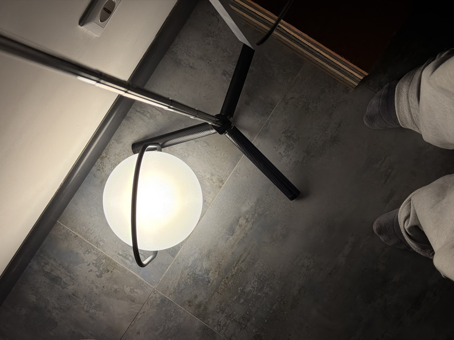 An indoor, high-angle shot captures a modern floor lamp and a person's lower legs and feet on a dark, textured floor. The perspective looks downwards, revealing the top-down view of the scene.
Dominating the left side of the frame is a contemporary floor lamp. Its primary light source is a spherical, frosted white glass diffuser, which emits a bright, warm glow, indicating it is turned on. This glowing orb is cradled within a minimalist black metal frame that wraps around it. The frame extends upwards to form part of the lamp's stand. The stand itself features three splayed legs made of a dark, possibly carbon fiber or ridged plastic, material, giving it a sturdy and modern appearance. A shiny, chrome-like pole extends vertically from the center of the stand, moving out of frame at the top.
The floor beneath the lamp and the person is composed of large, dark gray tiles or a concrete-like surface, exhibiting subtle variations in tone and texture that suggest a worn or industrial aesthetic. To the immediate left of the lamp, a white wall is visible, with a dark gray baseboard or trim running horizontally along its bottom edge. An electrical outlet, light gray or off-white in color with two horizontal slots and a grounding pin hole, is mounted on this wall near the top left corner of the image.
On the upper right side of the frame, a dark object with horizontal layers, possibly stacked wood or shelving, is partially visible, suggesting the presence of furniture. To the far right, a person's lower legs and feet are visible. The person is wearing light-colored (likely off-white or light gray) casual trousers or sweatpants, which are slightly bunched up at the ankles. Their feet are clad in dark gray socks with distinct lighter gray stripes around the ankle area.
The overall lighting suggests an indoor setting, possibly in the evening or a room with dim ambient light, where the lamp serves as a significant light source. No specific activity is depicted other than the presence of the lamp and the person. There is no visible text in the image.
