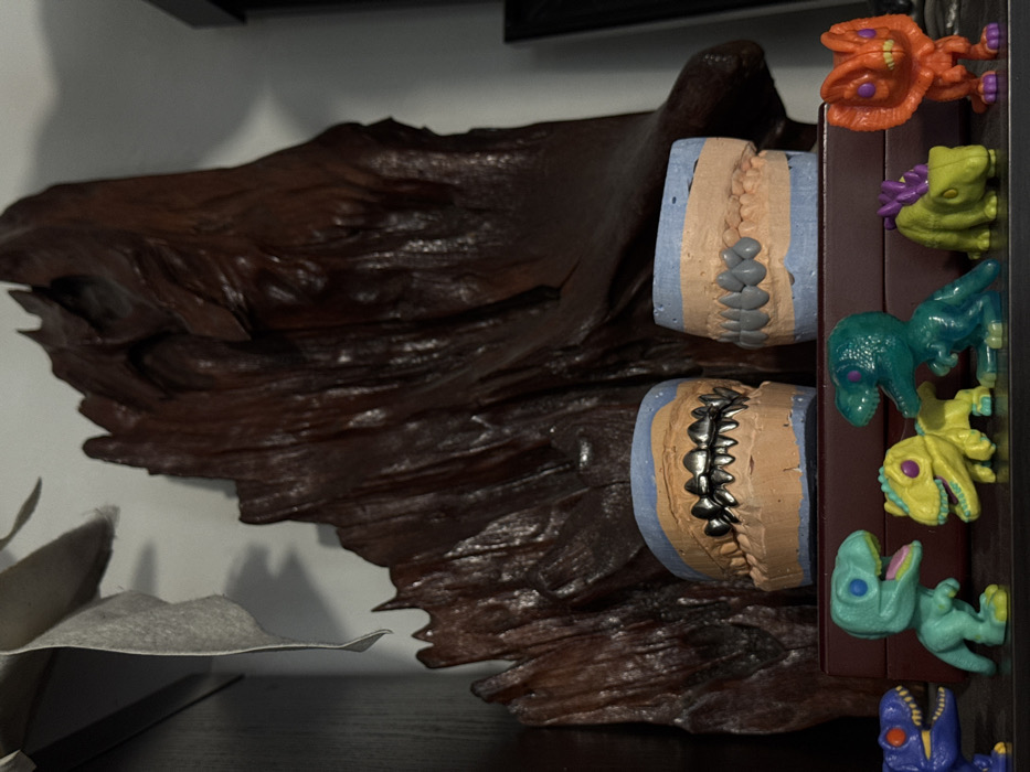 The scene depicts an indoor display in Sofia, Bulgaria. In the foreground, a dark horizontal surface features a row of six stylized dinosaur toys in various colors, including purple, teal, yellow, dark teal, lime green, and orange. Directly behind these toys, two dental models are positioned on a dark reddish-brown rectangular object. The left model, resting on a light blue base, presents peach-colored gums fitted with a complete set of silver-colored metal teeth. The right model, also on a light blue base, displays peach-colored gums with a full set of gray-colored teeth. Dominating the background is a large, dark brown, intricately textured wooden sculpture, possibly a root or stump, standing vertically against a light-colored wall. A piece of light-colored material hangs from the top left, casting a shadow on the wall. The overall environment suggests a curated display or workspace with ambient lighting.