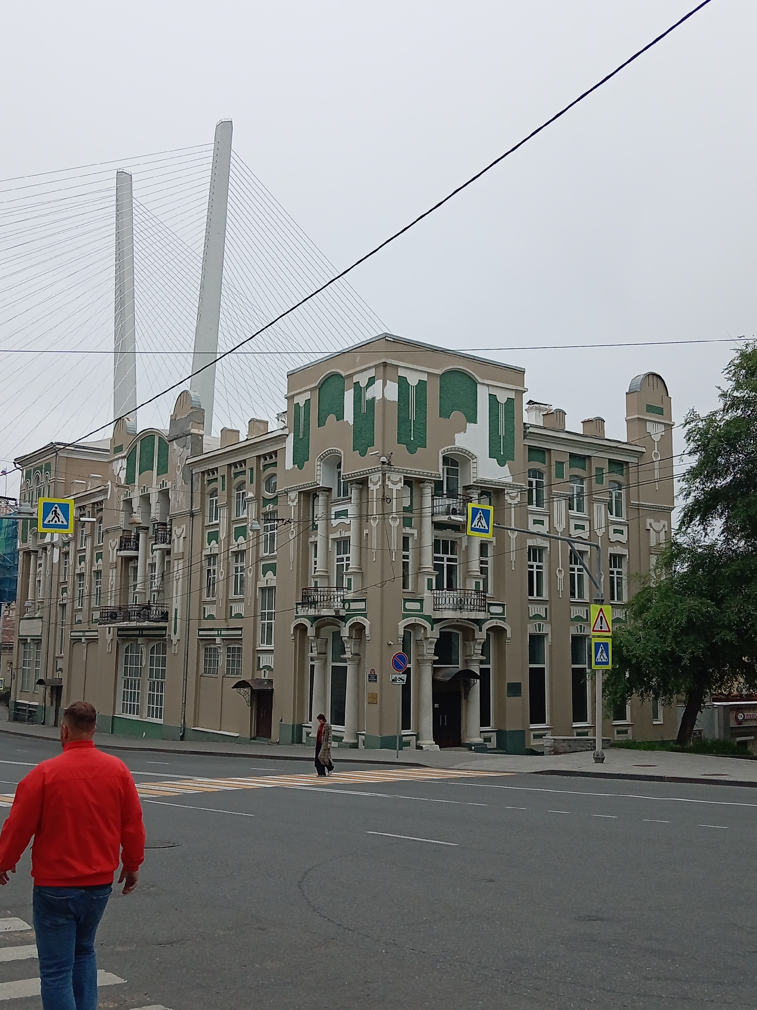 The image depicts a street scene in Vladivostok, Russia. In the foreground, a man wearing a red jacket and blue jeans is walking away from the camera, crossing a pedestrian crosswalk. Further in the background, another pedestrian, a woman wearing a patterned coat, is also crossing the street.
Dominating the mid-ground is an ornate, multi-story building with a beige facade and green decorative accents. The building features numerous windows, balconies with wrought-iron railings, and classical architectural elements such as columns. Several pedestrian crossing signs are visible on poles in front of the building.
In the background, the distinctive white towers and cable structure of the Golden Horn Bay Bridge (Russky Bridge) are visible against a cloudy sky. A large green tree is situated on the right side of the frame, partially obscuring the building. The street appears to be asphalted, with clear lane markings. Overhead, electrical wires crisscross the sky. The overall atmosphere is overcast.