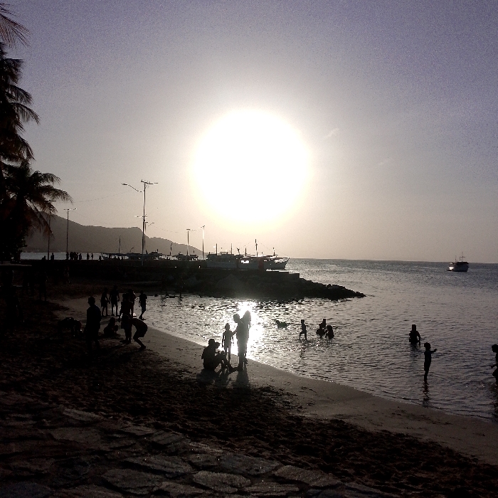 Silhouetted figures populate a beach at sunset. The sun, a brilliant orb, dominates the sky above the horizon line, reflecting on the water's surface. Several people are in the water while others stand or sit on the sandy beach. A jetty or breakwater extends into the sea, lined with boats and figures. Palm trees are visible on the left side, and in the distance, a land mass rises from the water. Light poles are visible on the coast line.