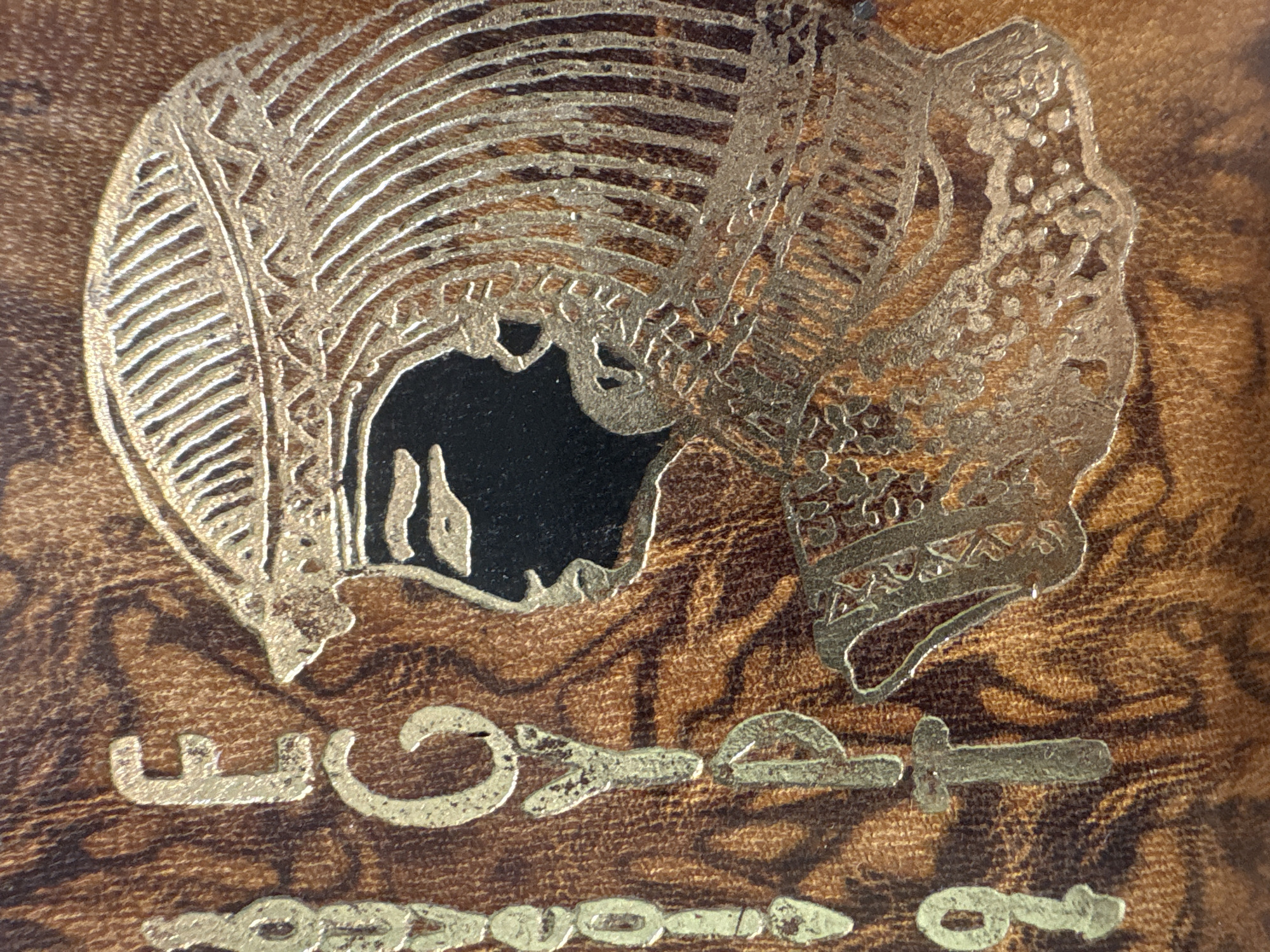 The image shows a close-up of what appears to be the cover of a book or perhaps a box. The cover is made of brown leather-like material with a textured grain. A design in gold/yellow metallic ink or foil is embossed onto the cover. This design features a stylized head (possibly a pharaoh or other figure from ancient Egypt) with elaborate headwear. Below the head are what appear to be hieroglyphs or symbolic characters. The image does not provide information about the location in the city of Krasnoyarsk, Russia.