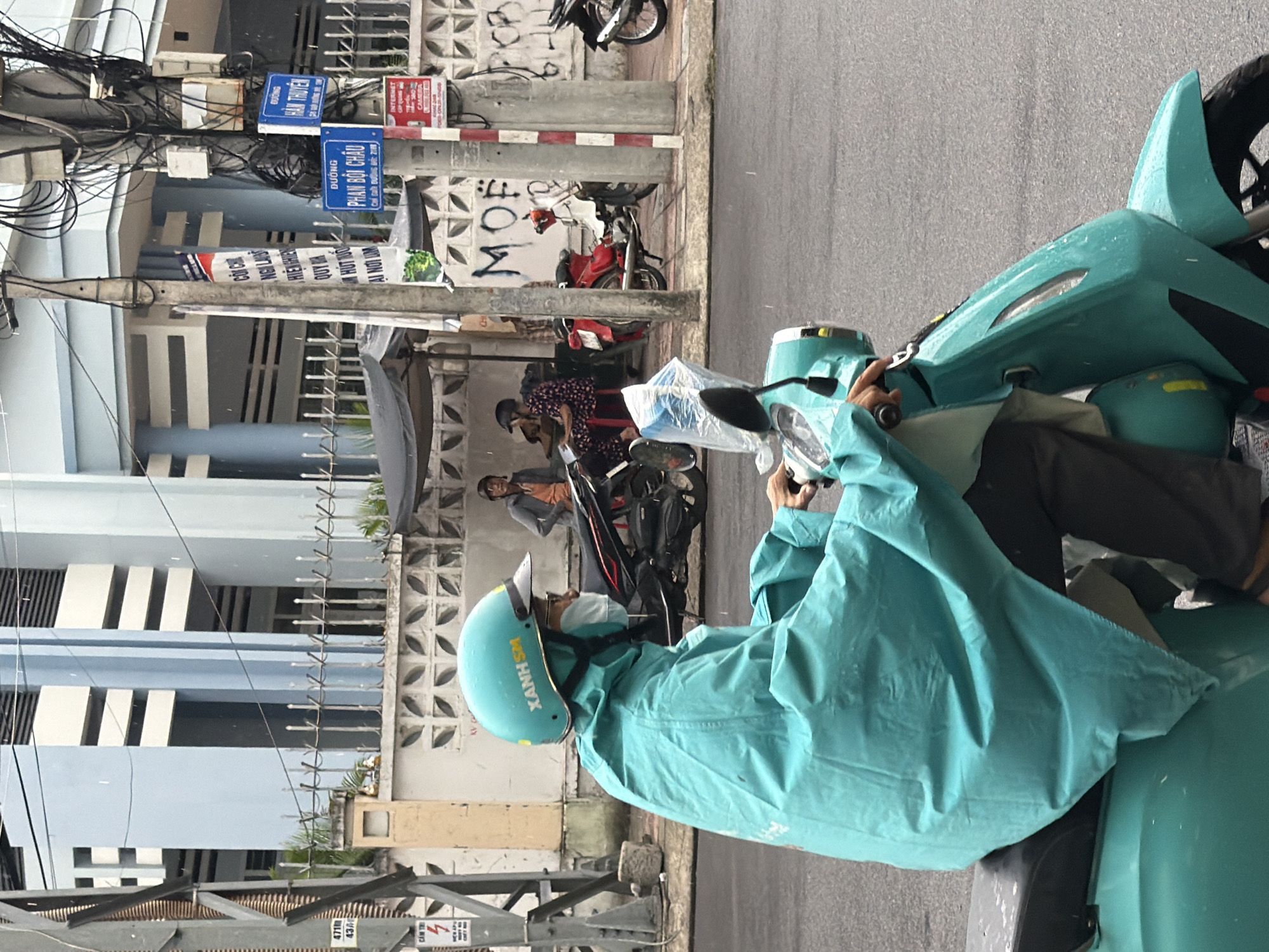 A person wearing a turquoise rain poncho and helmet rides a turquoise scooter on a wet asphalt road in Nha Trang, Vietnam. The person is wearing a white surgical mask. In the background, there is a building with light blue and white vertical accents, a grey concrete wall with decorative patterns, and a series of metal poles with wires and signs. One sign is a blue street sign that reads "PHAN BOI CHAU". Another blue sign reads "DUONG HAN THUYEN". Further in the background, a few other people are seen near a covered stall and parked motorcycles. It is raining, indicated by streaks of water in the air and on surfaces.