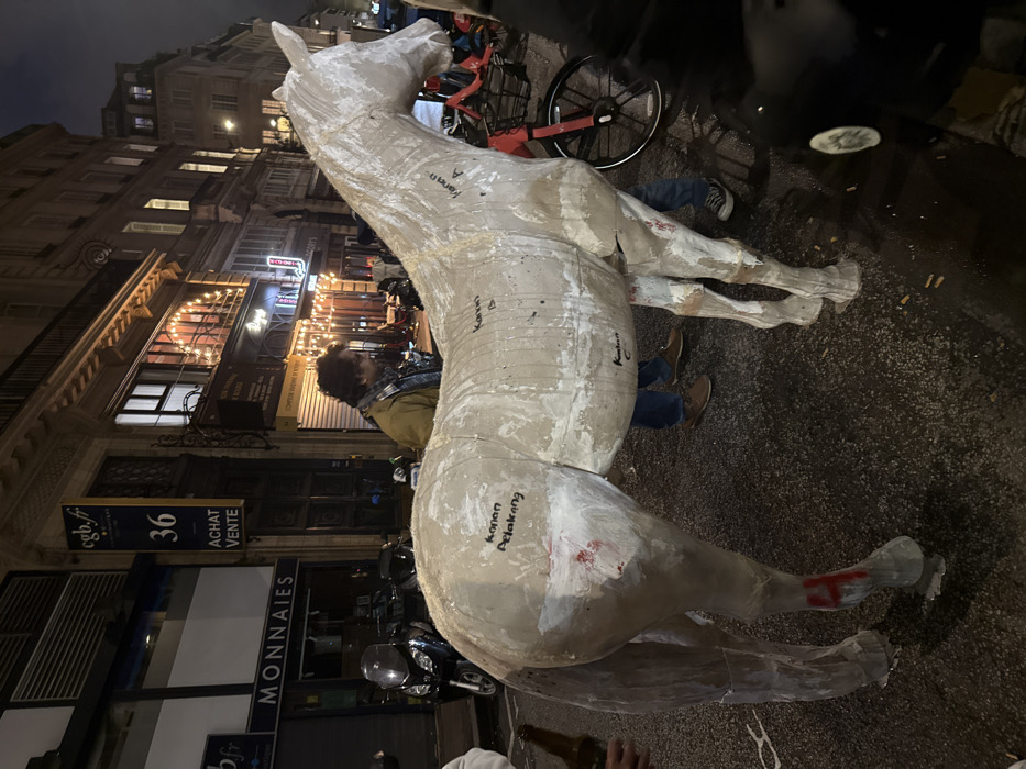 The image captures an evening street scene, likely in a European city, dominated by a large, light-colored horse statue. The overall impression is one of a public space after dark, illuminated by artificial lights, with a unique art object as the focal point.
The central subject is a life-sized or larger-than-life statue of a horse, made from a light-colored material such as plaster, paper-mache, or an unfinished composite. Its surface is textured and shows visible seams or layers, giving it a raw, unpolished, or artisanal appearance. The horse is standing upright, facing towards the upper-left of the frame. Handwritten labels in dark marker are visible on its body: "Kanan A" on the neck/shoulder, "Kanan C" on the belly, and "Kanan Belakang" on the hind leg/hip area. There's also some reddish mark or paint on its hind leg.
Several individuals are present around the horse. One person with dark, curly hair and a light-colored jacket is partially visible behind the horse's neck and shoulder, looking downwards. Another person's lower body, clad in blue jeans and brown shoes, is visible beneath the horse's belly, suggesting they are crouching or sitting close to the statue. At the very bottom edge of the frame, a hand is visible holding a brown glass bottle.
The horse statue is positioned on what appears to be a paved street or sidewalk. In the background, tall, multi-story buildings with classic European architectural details line the street. These buildings are brightly lit by numerous warm-toned artificial lights, including lights from windows, shopfronts, and decorative string lights on some facades, creating a vibrant evening ambiance.
Various objects are scattered or parked on the street. A red bicycle with black wheels is visible behind the horse. A black scooter or motorcycle is parked closer to the buildings. The ground itself shows some debris, including what appear to be cigarette butts and white splatters.
The dark sky and the pervasive artificial lighting confirm that the scene takes place at night or during late dusk.
Visible text provides clues about the location and businesses. On a vertical sign attached to one of the buildings, the following French text is clearly legible: "cgb.fr", "36", "ACHAT VENTE" (meaning "BUY SELL"), and "MONNAIES" (meaning "COINS" or "CURRENCY"). This indicates a shop specializing in numismatics or collectibles. Higher up, another partially visible sign displays "BYG".
The activity is not explicitly clear, but the presence of people around the marked horse statue, coupled with its seemingly unfinished state, suggests it might be an art installation, a prop for an event, or an object being prepared, moved, or displayed in a public urban setting. The casual postures of the people imply they are either observing, resting, or involved in setting up the statue.