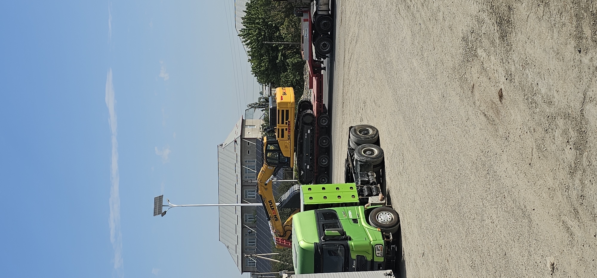 The image shows a construction site or transportation hub in Sheyhancha, Uzbekistan. In the foreground, a bright green semi-truck cab is parked on a gravel lot. Behind it, a yellow excavator is loaded onto a red flatbed trailer, which is itself being towed by another truck, partially visible on the right. The excavator has the brand name "SHANTUI" visible on its side. In the background, a long, grey, multi-story building with a distinctive gabled roof stands under a clear blue sky. A tall pole with a solar panel is positioned in front of the building, and other smaller structures and trees are visible. The ground is covered in gravel and dirt, with some tire tracks indicating recent vehicle activity.