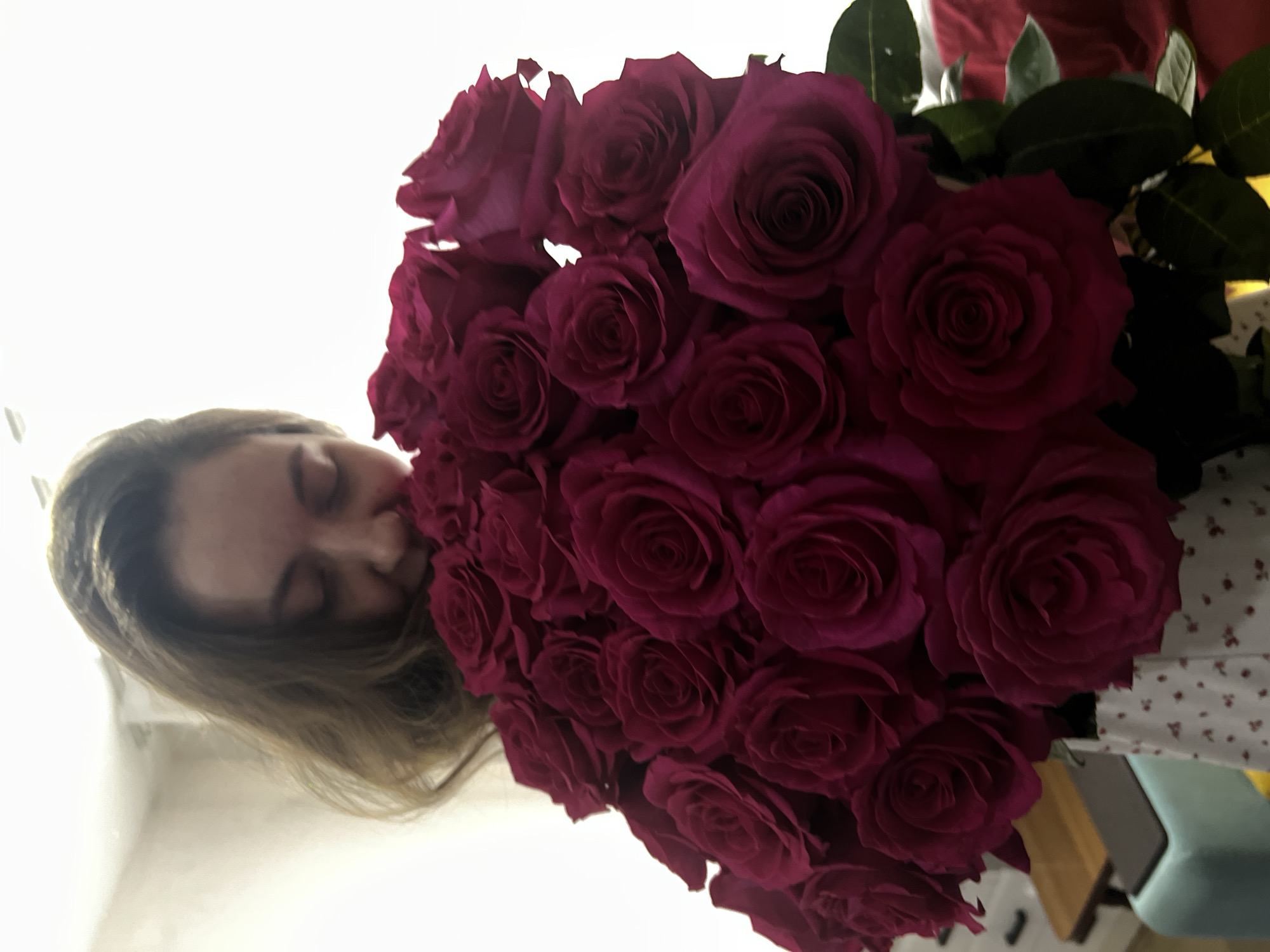 A woman with light brown hair is holding a large bouquet of dark pink roses. She is positioned to the left of the bouquet and appears to be smelling the roses. The background is blurry but seems to be a room with light-colored walls and some furniture. There is no specific information to confirm that this photo was taken in Votkinsk, Russia.