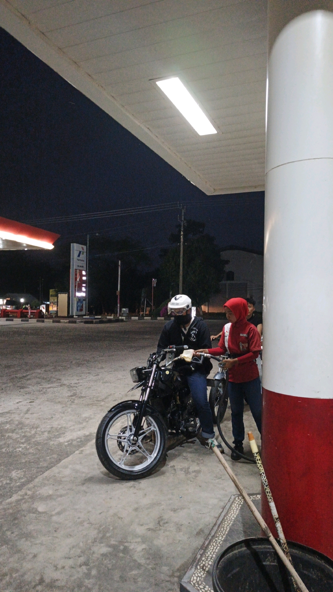At a gas station in Sukaresik, Indonesia, two individuals are depicted at night. A person wearing a white helmet and black jacket is seated on a black motorcycle, holding a fuel nozzle. Adjacent to them, a person in a red hijab and red shirt is also holding a fuel nozzle, appearing to be in the process of fueling the motorcycle. The environment is an outdoor gas station with a concrete ground, a white and red pillar, and a canopy overhead with fluorescent lighting. The sky is dark, indicating nighttime. In the background, there are signs for "PERTAMINA", a prominent Indonesian state-owned oil and gas company.