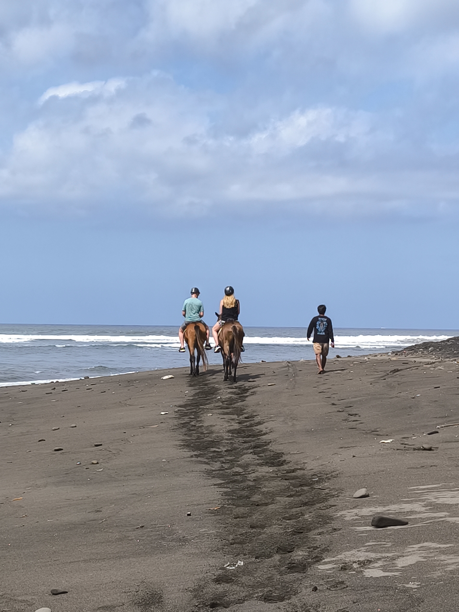 Two individuals on horseback ride along a dark sand beach adjacent to the ocean under a partly cloudy sky. A third individual walks on the beach behind them. The horses are brown, and the riders are wearing helmets. The person walking is wearing a dark long-sleeved shirt and shorts. The ocean has small waves. The beach is scattered with pebbles and debris. The image is taken from behind the subjects, facing the ocean.