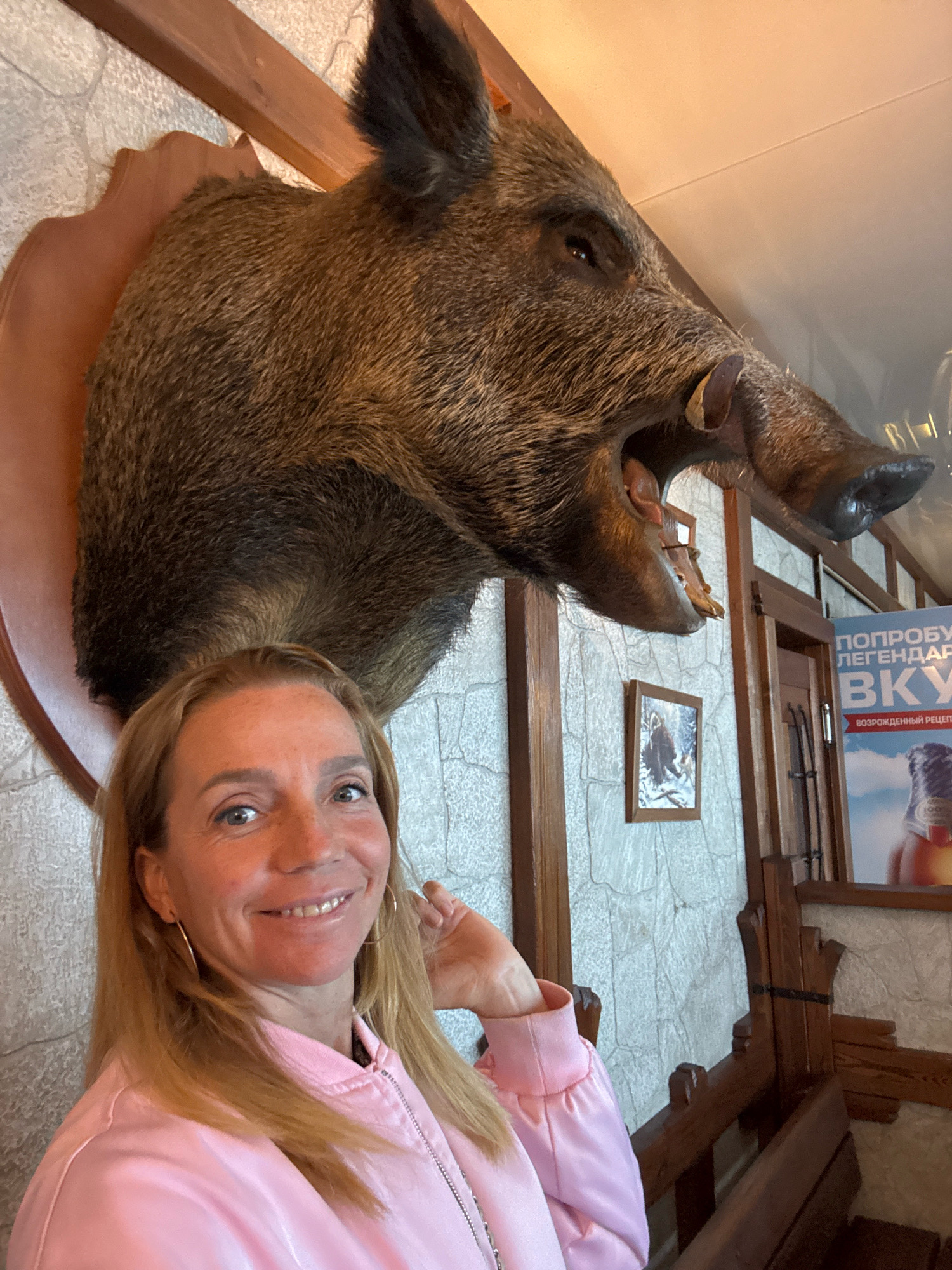 A woman with blonde hair and fair skin smiles at the camera, her head tilted slightly to the right. She is wearing a pink bomber jacket. Behind her, a taxidermied wild boar's head is mounted on a wooden plaque, its mouth open revealing tusks and teeth. The boar's head is positioned centrally in the upper portion of the image. The background consists of a stone-patterned wall, wooden paneling, and some framed pictures. To the right, a poster is visible, featuring text in Cyrillic script and an image of a bottle. The overall setting appears to be an indoor establishment, possibly a restaurant or a lodge, located in the city of Успенское, Russia.