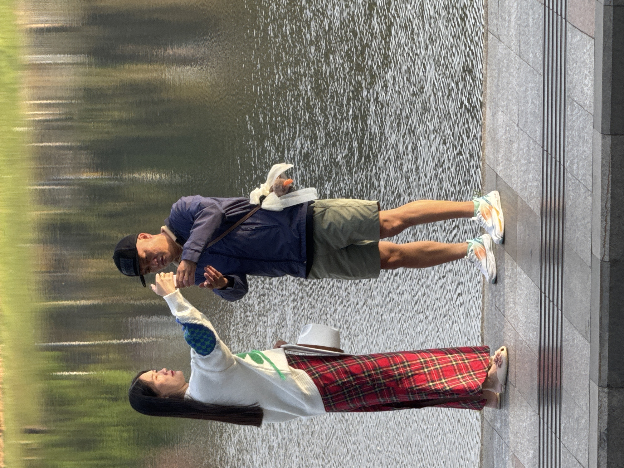 The image depicts two individuals standing on a paved walkway next to a body of water. The woman on the left, wearing a white sweater with green and blue checkered accents on the sleeves, a long red and black plaid skirt, and light-colored slip-on shoes, is holding a white hat. She is facing a man on her right. The man, dressed in a dark blue jacket, khaki shorts, a black baseball cap, and white athletic shoes, is holding out his right hand towards the woman. He appears to be receiving something from her, possibly a small white object. A white cloth bag with some contents is slung over his left shoulder. The water behind them reflects the green foliage of trees on the opposite bank, suggesting a park or natural setting. The paved walkway is composed of large gray tiles with a textured pattern. The overall lighting suggests it is either early morning or late afternoon, with sunlight casting shadows. This scene is consistent with the popular Xuan Huong Lake area in Da Lat, Vietnam, a well-known urban lake and park area that attracts locals and tourists.