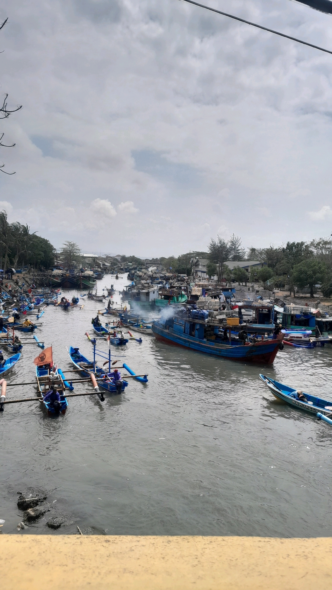 The image depicts a busy waterway in Kelurahan Tegalkamulyan, Cilacap, Indonesia, filled with numerous fishing boats of varying sizes. The water is murky and appears to be a river or canal. On the banks, a densely packed settlement with buildings and trees is visible. The sky is overcast with a mix of white and gray clouds. In the foreground, there is a yellow structure that seems to be a railing or bridge.
The waterway is crowded with boats, many of which are small, narrow fishing vessels with outriggers, some carrying one or two individuals. Larger, more robust fishing boats are also present, some emitting smoke, suggesting activity like engine operation or cooking. People are actively engaged in various tasks on and around the boats, including steering, preparing for departure, or tending to equipment. The density of boats and human activity indicates a bustling fishing port or harbor. The overall impression is one of a working waterfront community heavily reliant on maritime activities.