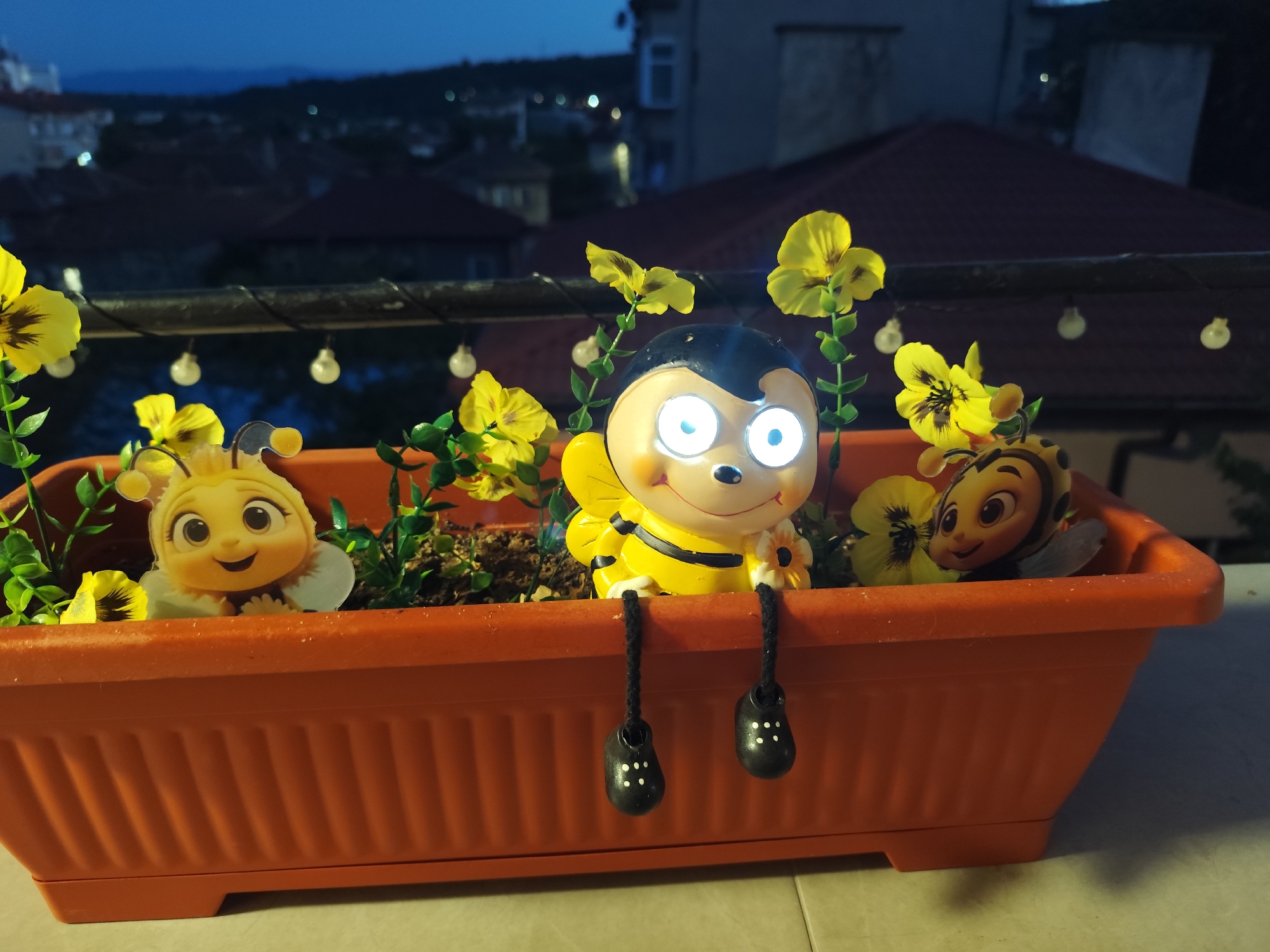 An orange rectangular planter sits on a light-colored tiled surface. The planter contains artificial yellow flowers and green foliage. Three bee-themed decorative figures are placed within the planter. The leftmost figure is a stylized bee character with a smiling face and large eyes, appearing as a cutout. In the center is a larger, three-dimensional bee figure with a black head, large illuminated eyes, yellow and black striped body, and black dangling feet with white dots. To the right is another cutout bee character with large eyes and a smiling mouth. The planter is positioned on a surface with a blurred background showing a cityscape at dusk or night. Lights from buildings are visible in the distance, along with rooftops and trees. A dark railing with string lights is in front of the planter. The overall scene appears to be outdoors, possibly on a balcony or patio, overlooking a town in Montana, Bulgaria.