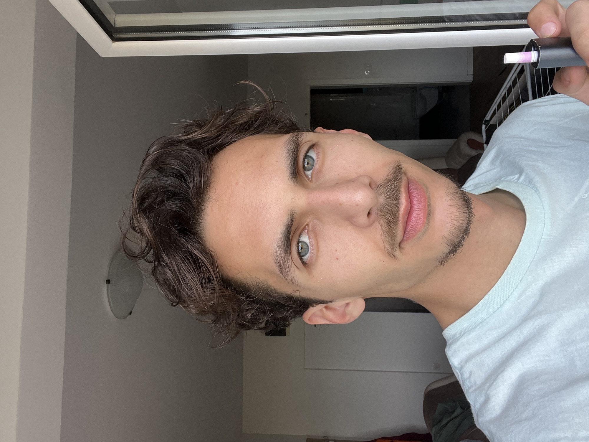 A man is holding a gray smoking device with a white cigarette inserted, facing the camera at an angle. He has curly brown hair, light blue eyes, and a mustache and goatee. He is wearing a light blue T-shirt. A window is visible behind him, and what appears to be the interior of an apartment or building, including a light fixture on the wall. There is no clear indication that this image was taken in the city of Rafailovići, Montenegro.