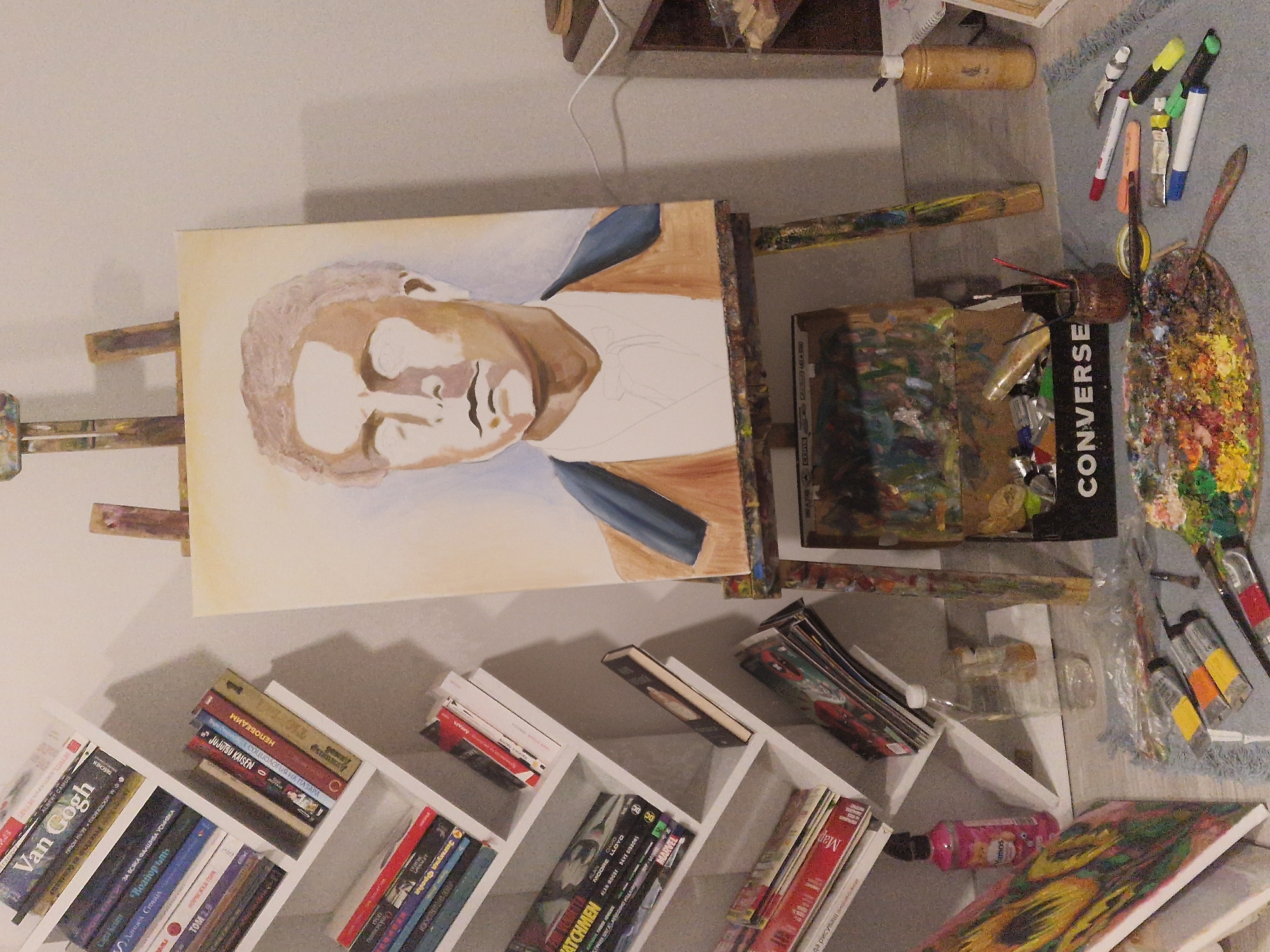 Here's a detailed description of the image:
**Scene:** An artist's workspace in an indoor setting.
**Objects:**
* **Artwork:** There's an unfinished portrait painting on an easel, depicting a person with brown skin tones and what appears to be a blue shirt or jacket. Another painting depicting a sunflower is present.
* **Bookshelf:** A white, asymmetrical bookshelf is filled with books, including volumes by "Van Gogh", "Albert Camus", a book in Bulgarian titled "За Всяка Фалшива Усмивка" ("For Every False Smile"), "Jujutsu Kaisen", "Watchmen", and a "Marvel" book, among others.
* **Art Supplies:** Various art supplies are scattered around the scene, including paint tubes, brushes, a palette with colorful paints, and a container of brushes in a Converse shoe box.
* **Other:** A rug, a wooden shelf, bottles, and a paint container are also visible.
**People:** The primary subject is the unfinished portrait of a person.
**Subjects:** Artwork, books, art supplies.
**Environment:** The lighting appears to be soft and diffused. The space looks cluttered, suggesting an active working environment.