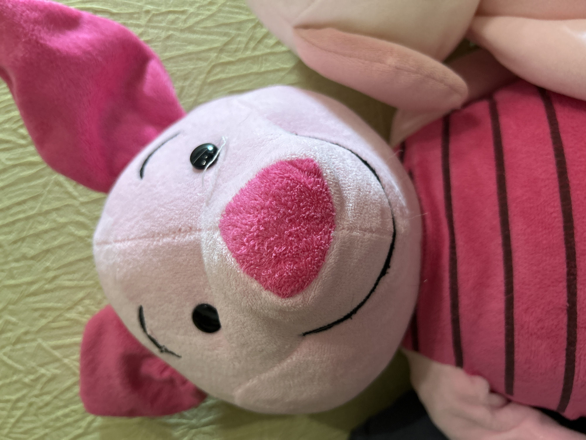 A close-up, eye-level shot shows a pink stuffed pig with large pink ears and a pink snout with a textured surface. The pig has black oval eyes and a black curved smile. It is wearing a pink shirt with horizontal dark brown stripes. To the right of the pig, another pink stuffed animal's ear and arm are visible. The background is a textured greenish-yellow surface, possibly a fabric or wall. The image is taken indoors, with no discernible connection to Moscow, Russia.