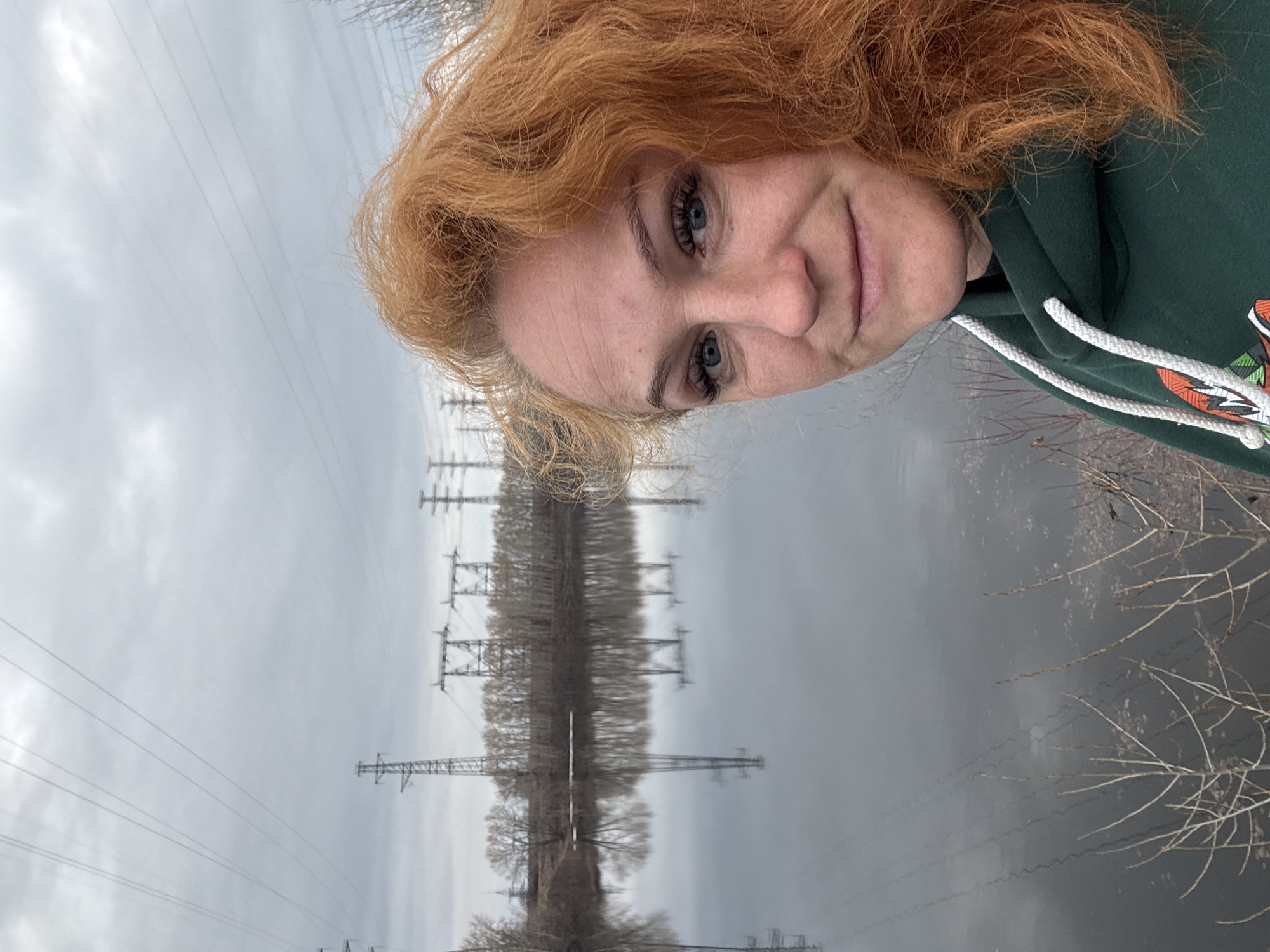 A photograph shows a red-haired person with blue eyes wearing a green hoodie with white drawstrings in the city of Chaikovsky, Russia. In the background, power lines and bare trees are reflected in a body of water. The sky is overcast.