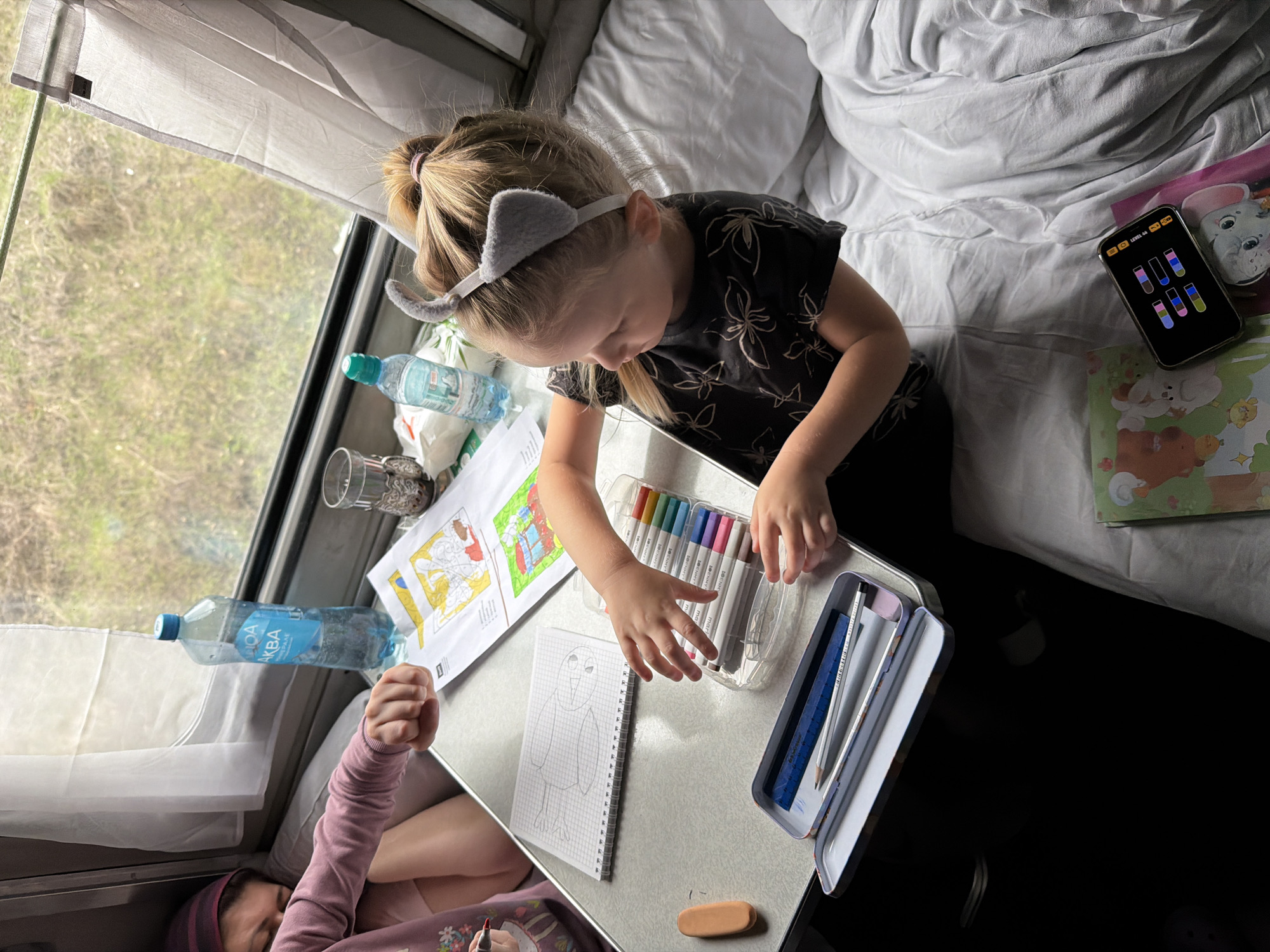 A young girl in a black flower-patterned shirt and gray cat-ear headband sits at a small table, drawing on a notebook. A woman in a pink sweater lies beneath the table. The table holds a box of markers, a pencil case with a ruler and pencil, and water bottles. Coloring books and a cell phone displaying a game are on the bed above. The background shows a window with blurred scenery. The location is not specified, and there is no evidence to suggest the image was taken in the city of Korovino, Russia.