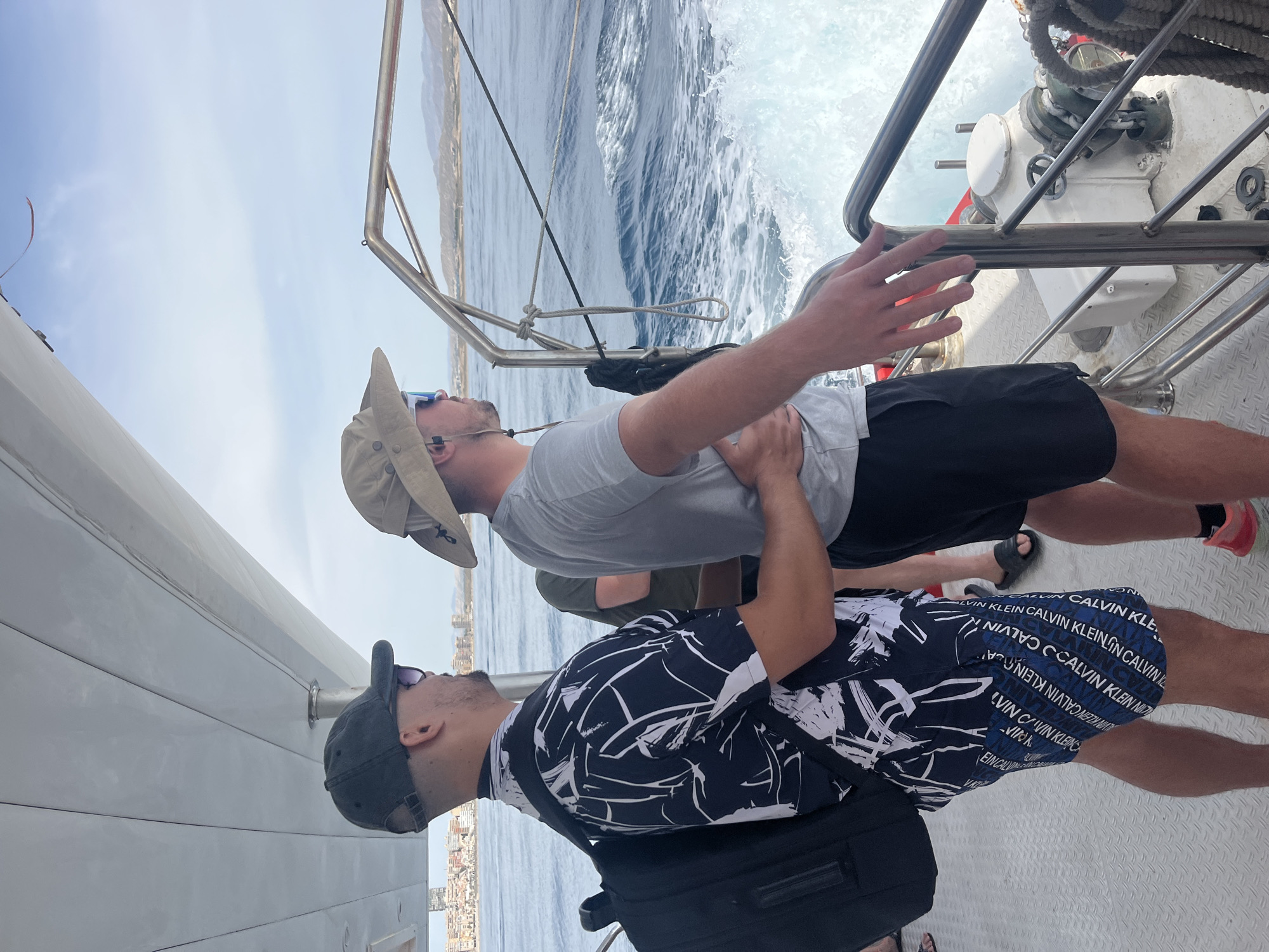 Two men are standing on the deck of a boat, looking out at the ocean. The man on the left is wearing a black and white patterned shirt, blue and white patterned shorts, a dark baseball cap, and sunglasses. He has a black backpack on his back. The man on the right is wearing a light grey t-shirt, black shorts, a tan bucket hat, and sunglasses. He has his left arm around the waist of the man on the left. The boat is moving, as evidenced by the wake behind it. In the distance, a city skyline is visible along the coast. The sky is blue with some clouds. The scene appears to be taking place in the Mediterranean Sea, possibly near Elx/Elche, Spain, given the architecture of the distant city.