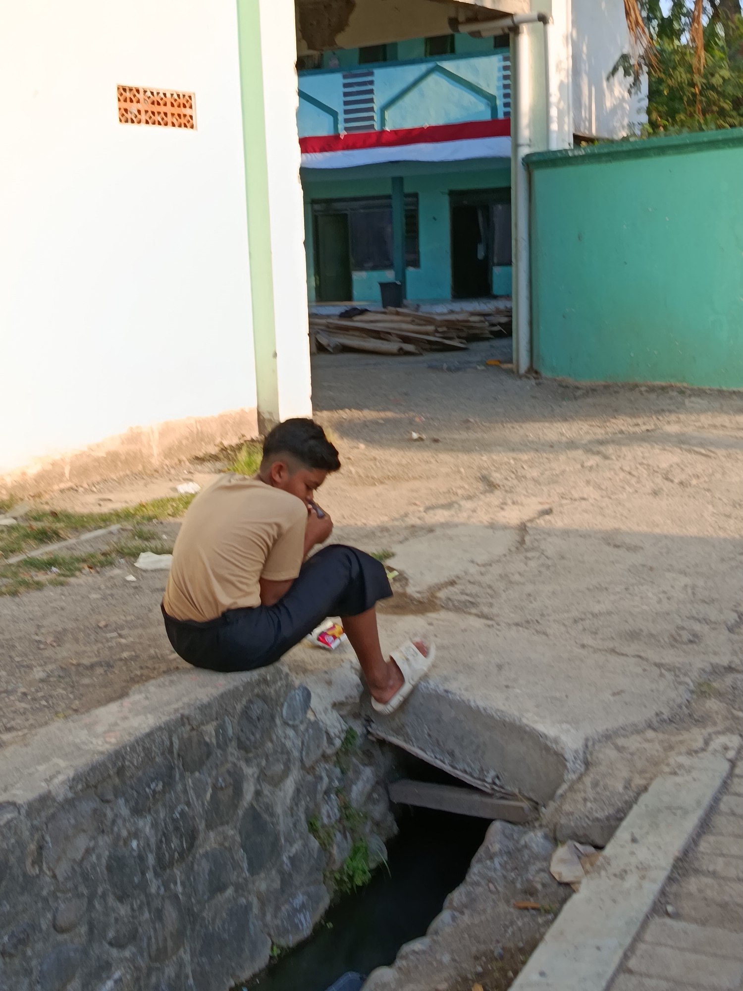 A young boy is seated on a concrete ledge next to a drainage ditch. He is wearing a tan t-shirt and dark pants, with white sandals on his feet. The boy is holding a small object to his mouth, and appears to be looking down at it. Behind him, a building with light blue and white sections is visible. The building is decorated with a red and white flag, which may be the Indonesian flag. The ground in front of the building is covered in sand and gravel, and there are stacks of wood. The scene appears to be in a semi-urban or rural area, likely in Ramban Wetan, Indonesia, given the style of the building and the presence of what appears to be the Indonesian flag.