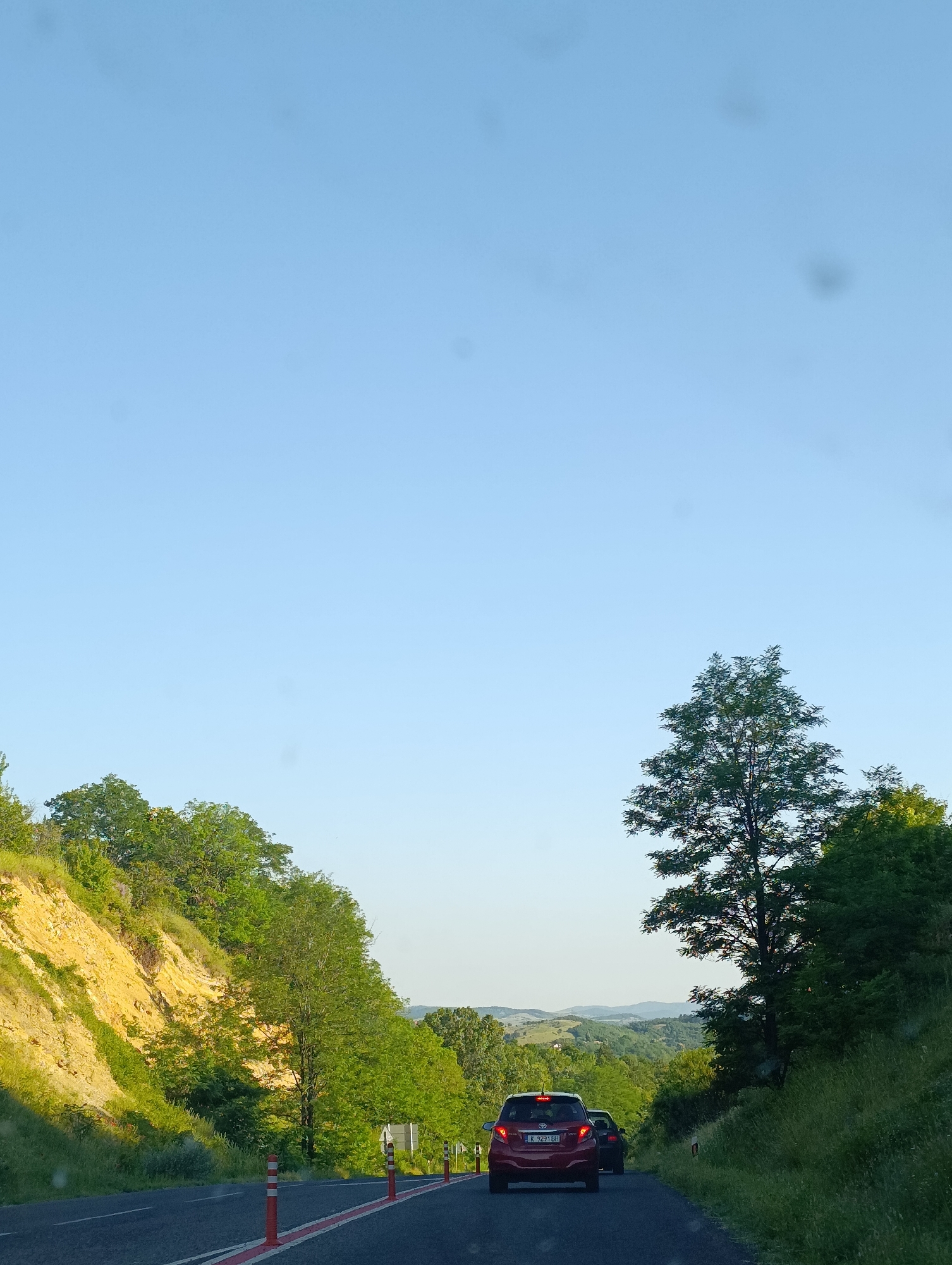 The image depicts a view from a moving vehicle on a road. The road is asphalt with a red and white line marking the lane. To the left of the road, a steep, yellowish-brown embankment is covered in green vegetation. To the right, lush green grass and trees line the roadside. In the distance, a rolling landscape of green hills under a clear blue sky is visible.
Two cars are on the road ahead. The closest car is a red hatchback, its rear facing the camera. It has a license plate that appears to read "K 9291 BH". Behind the red car, a darker colored vehicle is partially visible.
Several orange and white traffic bollards are placed along the edge of the road. The scene suggests a rural or semi-rural setting, possibly along a scenic route in Bulgaria, given the context of Cherna skala. The lighting indicates it is likely daytime, with the sun casting shadows. There are some blurry spots on the image, likely from dirt or water droplets on the camera lens.