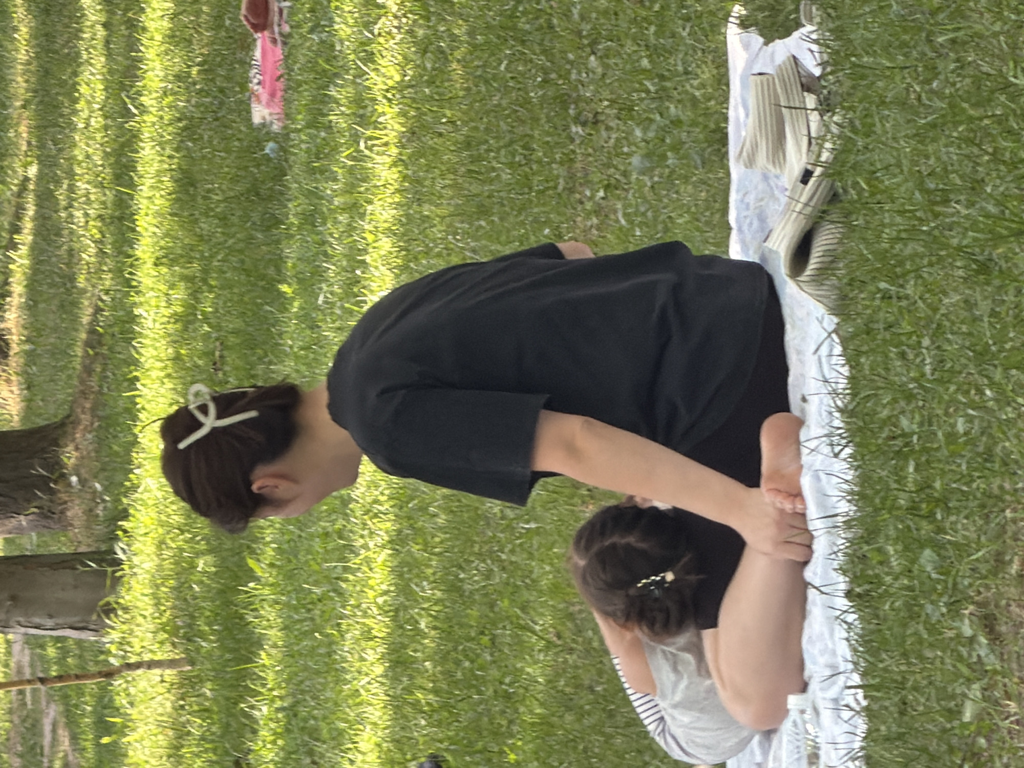 The image depicts two individuals on a grassy area, possibly a park. A woman with dark hair tied back with a white clip is seated facing away from the camera, wearing a black t-shirt. In front of her, a child with dark hair styled in a braid and a small white clip is lying on their side on a white blanket, their head bent forward. The child is wearing a striped shirt. A folded hat with a striped pattern rests on the blanket next to the child. The background consists of green grass, trees, and dappled sunlight. The overall scene suggests a relaxed outdoor activity, such as a picnic or yoga session.