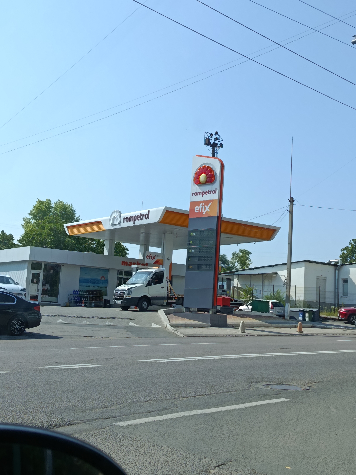 The image depicts a Rompetrol gas station in Chișinău, Moldova, on a clear, sunny day. A tall, illuminated sign featuring the Rompetrol logo (a stylized flower) and fuel prices stands prominently. The station itself has a white canopy with orange trim, under which several fuel pumps are visible. A white tow truck with its hazard lights on is parked near the pumps. In front of the station is a market building with large windows displaying various products, including bottles of oil. Several cars are present, including a black sedan in the foreground and a white van. The scene is situated along a road with visible lane markings and a manhole cover. Trees and a few other buildings are visible in the background, suggesting an urban or suburban setting. The sky is a clear blue with visible power lines stretching across it.