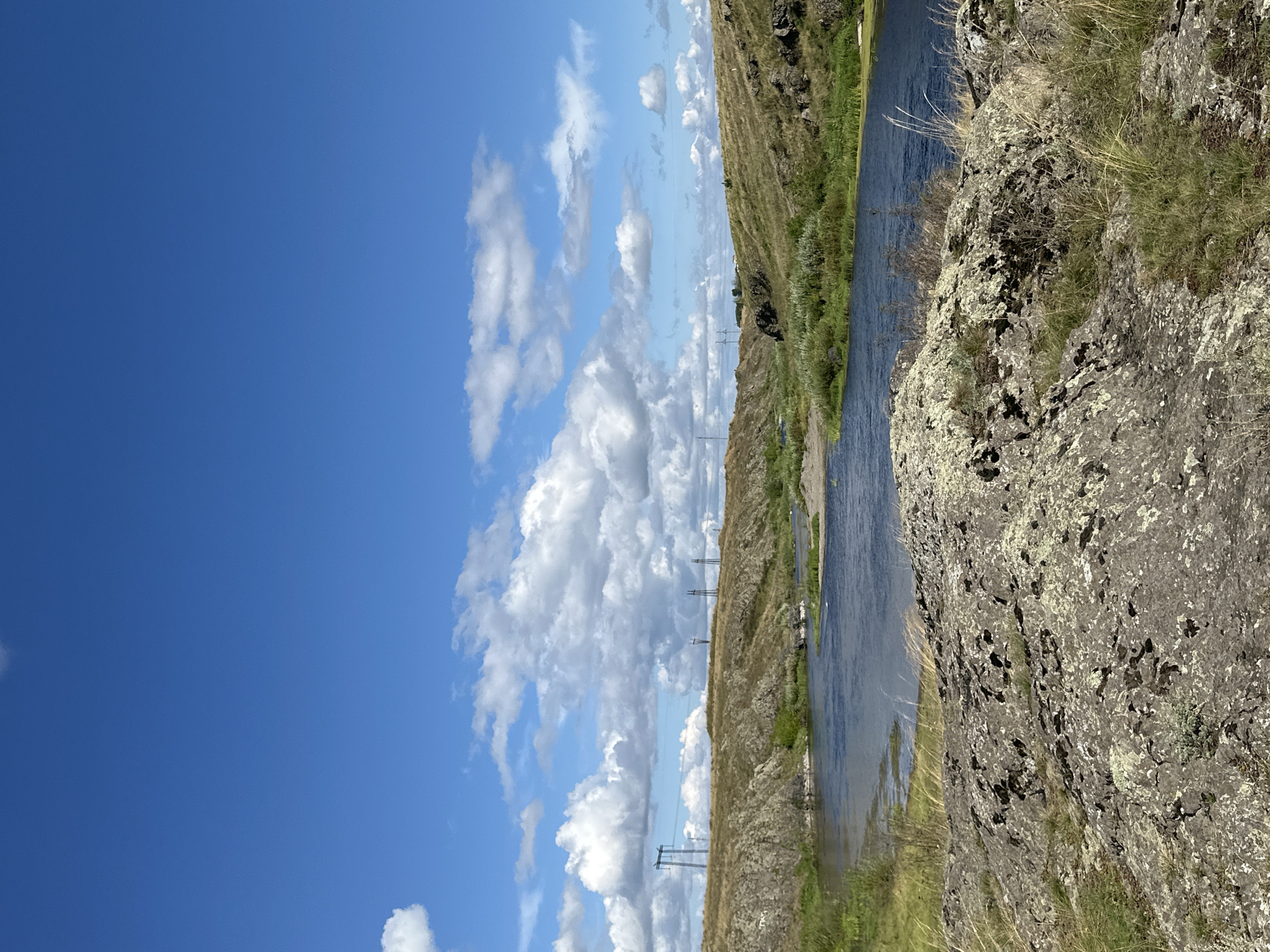 A wide shot captures a natural landscape under a partly cloudy blue sky. In the foreground, large, textured rocks covered in lichen are visible, with dry grass growing around them. A body of water, likely a river, flows through the middle ground, its surface reflecting the sky. The riverbanks are lined with dry, golden grass and patches of green vegetation. In the background, rolling hills covered in dry grass and rocky outcrops rise up. Several tall, industrial power line towers with electrical wires strung between them are scattered across the hills, indicating a human presence in this otherwise natural setting. The overall impression is one of a remote, open landscape, possibly a valley or a gorge.