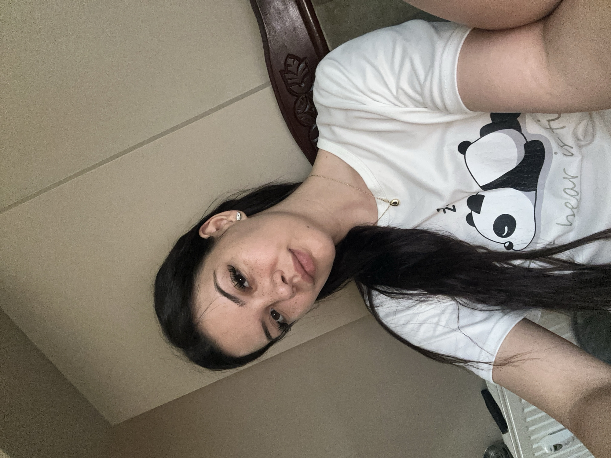 In Sliven, Bulgaria, a young woman with dark hair and freckles is seen in a selfie. She is wearing a white t-shirt with a panda graphic and the text "bear is ti". A gold necklace with a dark pendant is visible. The background consists of a beige wall with a subtle texture and a dark wooden headboard.
