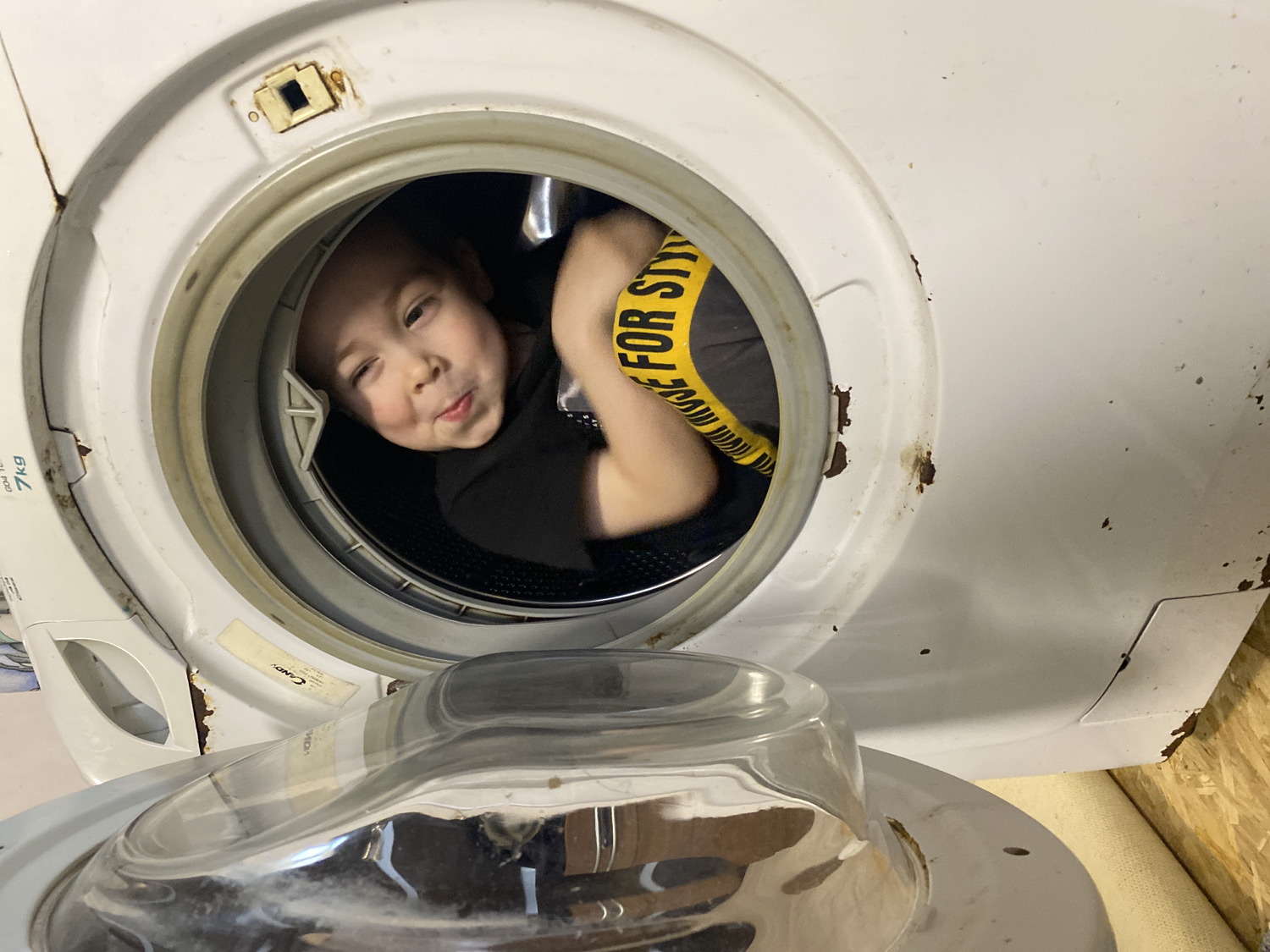 This indoor photograph, taken in Noviy, Russia, captures a young child playfully positioned inside the drum of an old, front-loading washing machine. The child, likely a boy, is seen from the chest up, smiling mischievously at the camera with a playful smirk. He is wearing a black t-shirt, and a yellow and black striped strap or piece of clothing with partially visible text "FOR STY" (likely "FOR STYLE") is visible on his right arm/chest.
The washing machine itself is a prominent feature, showing significant signs of age and wear. Its white exterior is discolored and covered with numerous rust spots, particularly around the circular opening where the child is located, and along the edges of the machine's body. The brand name "CANDY" is visible on a label inside the door rim, along with "7kg" indicating its capacity. The front-loading door is open and swung downwards, with its transparent, domed cover reflecting some light. The interior of the drum is dark, contrasting with the child's bright face. The immediate surroundings suggest a utilitarian space, possibly a laundry area, with a wooden surface visible on the right. The overall mood is lighthearted and evokes childhood curiosity and playfulness in an ordinary domestic setting.