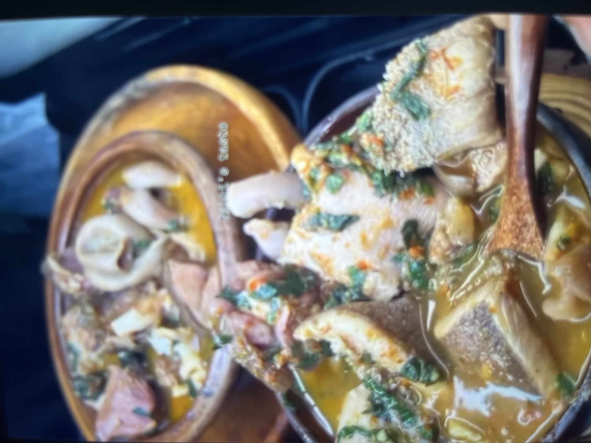 The image displays two clay bowls filled with a hearty stew. The stew appears to be a rich, yellow broth containing various meats and vegetables, generously garnished with chopped green herbs and red pepper flakes. The texture of the meat suggests it includes tripe and possibly other offal, with some pieces having a gelatinous appearance. One bowl, positioned in the foreground, is partially obscured by a wooden spoon. The second bowl sits behind it, with a wooden serving plate beneath it. The overall presentation is rustic and appetizing, indicative of traditional African cuisine. The scene is filmed indoors, with a dark background that suggests a home or restaurant setting. There are no discernible landmarks or environmental cues that would pinpoint the location to Abuja, Nigeria. The text "Mama's taste" is visible in the upper portion of the image, superimposed over the food.