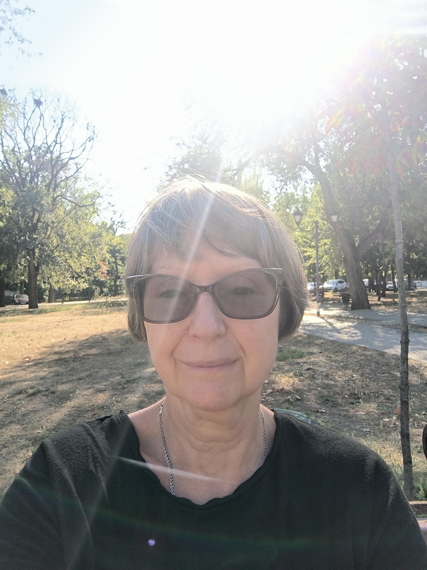 This is a selfie taken outdoors in a park-like setting with trees and dry grass on a sunny day. The sun is bright and directly overhead, causing lens flare. The subject is a middle-aged Caucasian woman with short, grey hair, wearing sunglasses and a black t-shirt. She is smiling. In the background, there are more trees, a paved pathway, streetlights, and parked cars. The location is the city of Mykolaiv, Ukraine.