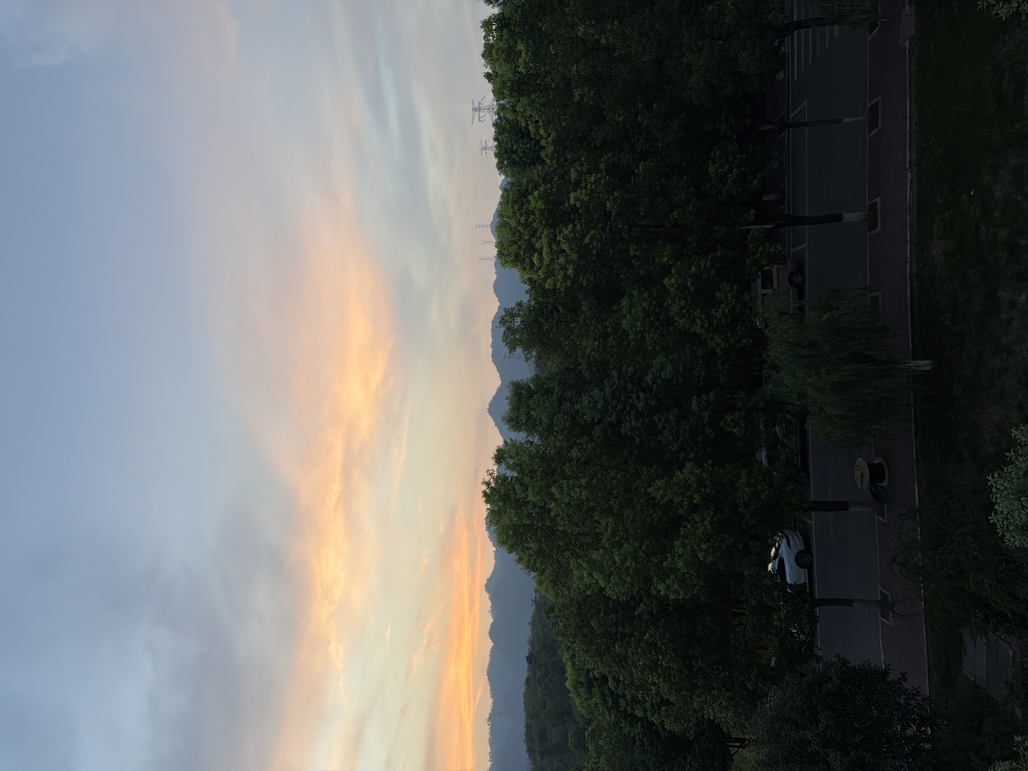 This is a high-angle, wide shot of a tree-lined street during dusk in Jiangjin, China. The sky is a gradient of soft blues and pinks, with wispy clouds catching the last light of the setting sun. In the distance, rolling hills are silhouetted against the colorful sky, with several power transmission towers visible on the horizon.
In the foreground and midground, a dense canopy of lush green trees forms a natural border along the street. The street itself is paved, with a brick walkway bordering one side. A white car is parked on the side of the road, its reflection shimmering in the surface. Further down the street, another vehicle is partially visible. A spool of wire or cable rests on the walkway. The overall impression is one of a serene, semi-urban landscape at the close of day.
