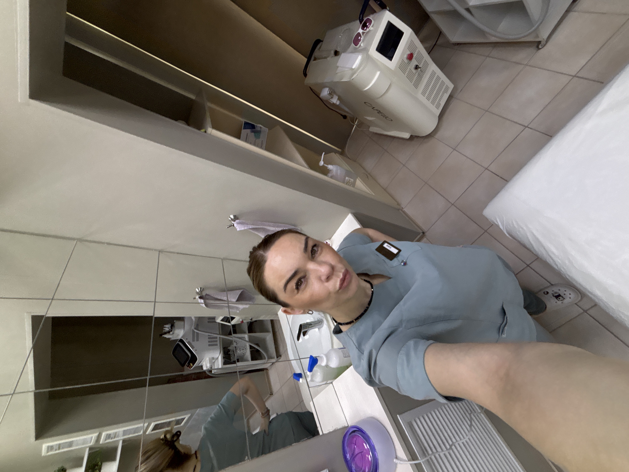 The image shows a person taking a selfie in what appears to be a medical or beauty treatment room. The room includes a white medical bed, a cosmetic laser machine, a sink, and shelves with various items. The person taking the selfie is wearing light blue scrubs and a black choker. The reflection in a tiled wall shows the room's contents and another angle of the person. The location is suggested to be Vsevolozhsk, Russia.