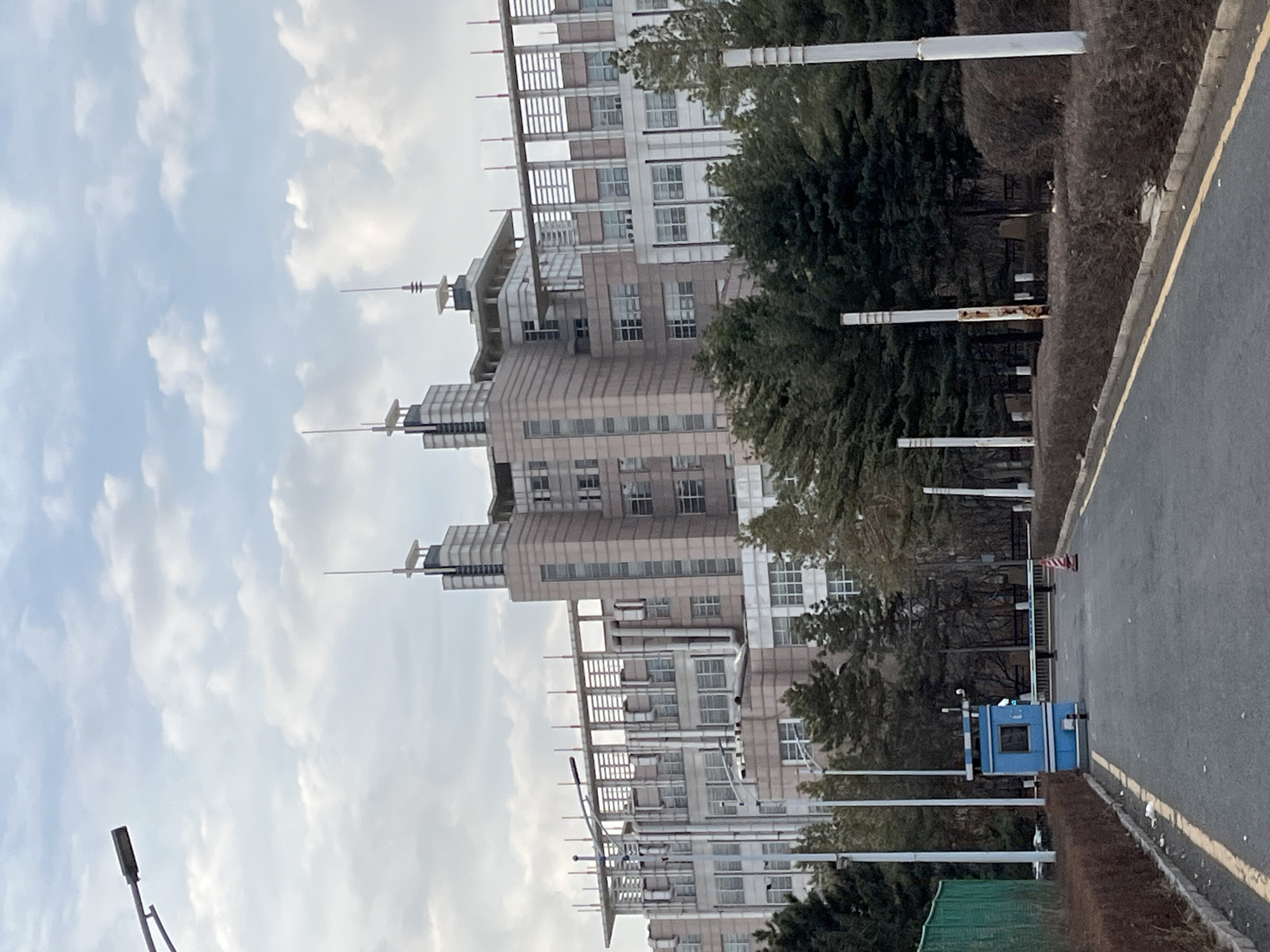 The image shows a multi-story building in Changchun City, China. The building has a layered design with multiple setbacks. The exterior is a light brown color with numerous windows. Tall antennas are affixed to the top of the building. Along the street adjacent to the building are trees, shrubs, and a blue security booth. A gray asphalt road runs along the right side of the frame. The sky is overcast with scattered clouds. The orientation of the photograph is sideways.