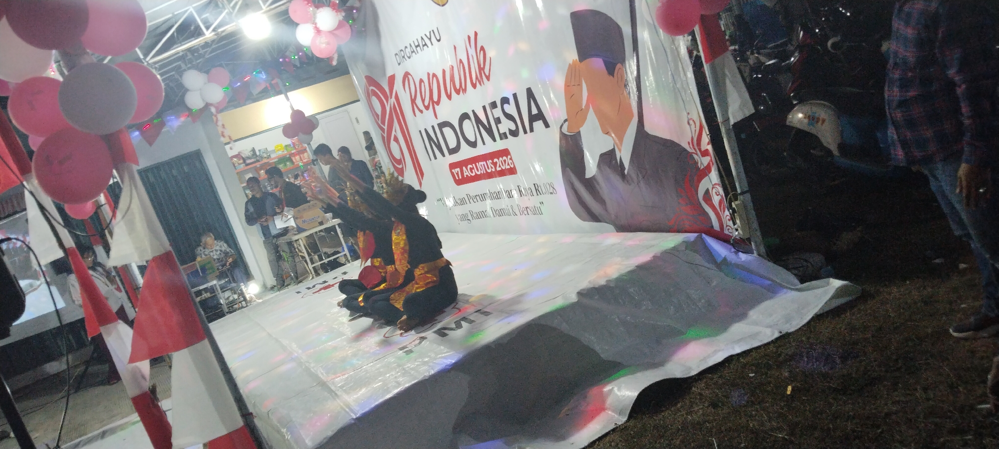 This image depicts an outdoor event, possibly a celebration or performance, with a large banner featuring a stylized depiction of a man in a suit and hat, saluting, superimposed over text reading "DIRGAHAYU REPUBLIK INDONESIA 17 AGUSTUS 2026". Balloons in red, white, and pink are visible at the top of the frame, as are Indonesian flags. In the foreground, two individuals dressed in traditional Indonesian attire, with golden sashes and headpieces, are seated on a white tarp, performing a dance or ritual. The background shows a tent-like structure, some tables with people gathered around them, and motorcycles parked in the darker area to the right. The ground appears to be grass. The overall scene suggests a community event celebrating Indonesian Independence Day. The location is identified as Taban, Indonesia.