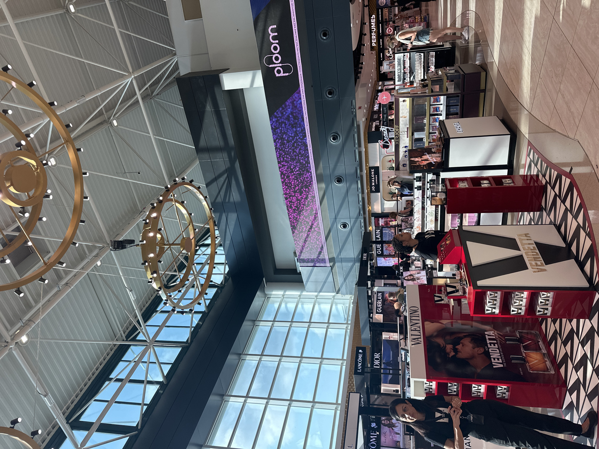 The image depicts a retail environment, likely an airport duty-free shop, characterized by an expansive interior with a high ceiling featuring modern industrial architecture and large windows. Two prominent, circular golden chandeliers with multiple lights hang from the ceiling. The lower level is dominated by displays of perfumes and cosmetics from various brands, including Valentino, Dior, Lancôme, and Guerlain. In the foreground, two female employees wearing black t-shirts with a "V" logo stand near a Valentino "Vendetta" perfume display. One employee, on the left, is looking away from the camera. The other employee, on the right, is looking towards the right side of the frame. Several customers are visible browsing the merchandise in the background. A large banner advertising "Ploom" is displayed on an upper wall. The flooring consists of patterned tiles and polished concrete, with sunlight streaming in from the windows. The scene suggests a busy commercial setting.