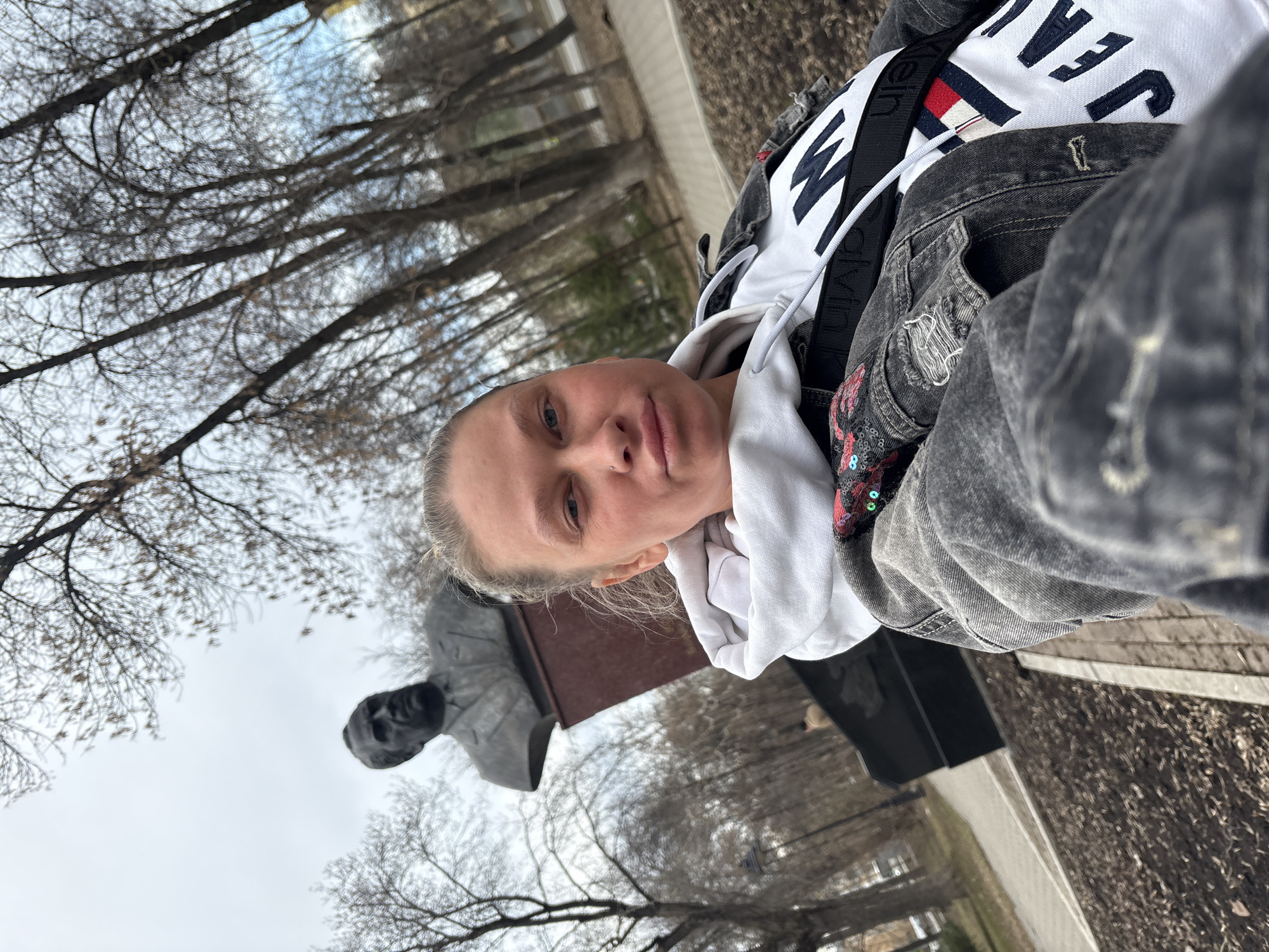 A selfie of a woman outdoors on a cloudy day. She is standing next to a bust of Aleksandr Stepanovich Popov, a Russian physicist and electrical engineer, located in the city of Perm, Russia. The woman is wearing a white hoodie, a black denim jacket with decorative sequins, and a Calvin Klein cross-body bag. The statue sits atop a dark base, near a sidewalk. Leafless trees are visible in the background.