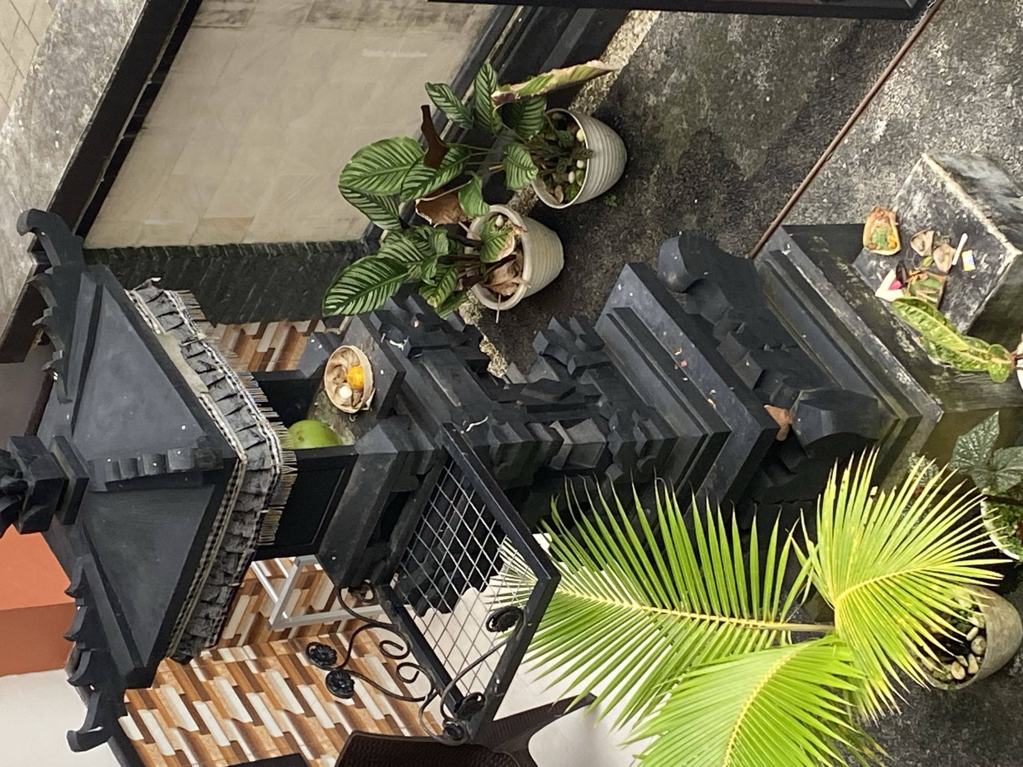 This eye-level shot, taken in Nusa Dua, Indonesia, depicts an outdoor arrangement of plants and a black shrine-like structure. Two potted plants with patterned leaves sit atop the shrine. The shrine itself is multi-tiered and has a woven lattice accent. Inside, a bowl of offerings rests on one tier. A potted palm tree dominates the foreground, its fronds arching. The background features a mix of tiled flooring and a dark, textured surface.