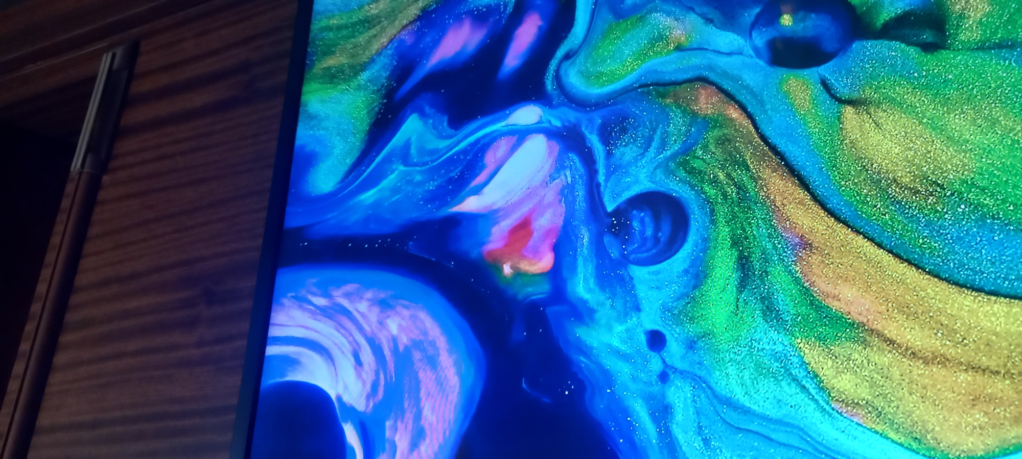 The image displays a close-up, abstract view of what appears to be a liquid art or marbling effect on a screen. The dominant colors are vibrant blues, greens, and purples, with streaks of pink and orange. There are areas of glittery texture, particularly in the green and gold sections. The composition is dynamic, with swirling patterns and fluid shapes. A dark, circular object with a reflective surface is visible in the lower right corner. The top of the image shows a portion of a wooden surface, possibly a desk or shelf, with a thin metallic bar.
There is no indication or information within the image to suggest its location in the city of Kryvyi Rih, Ukraine.