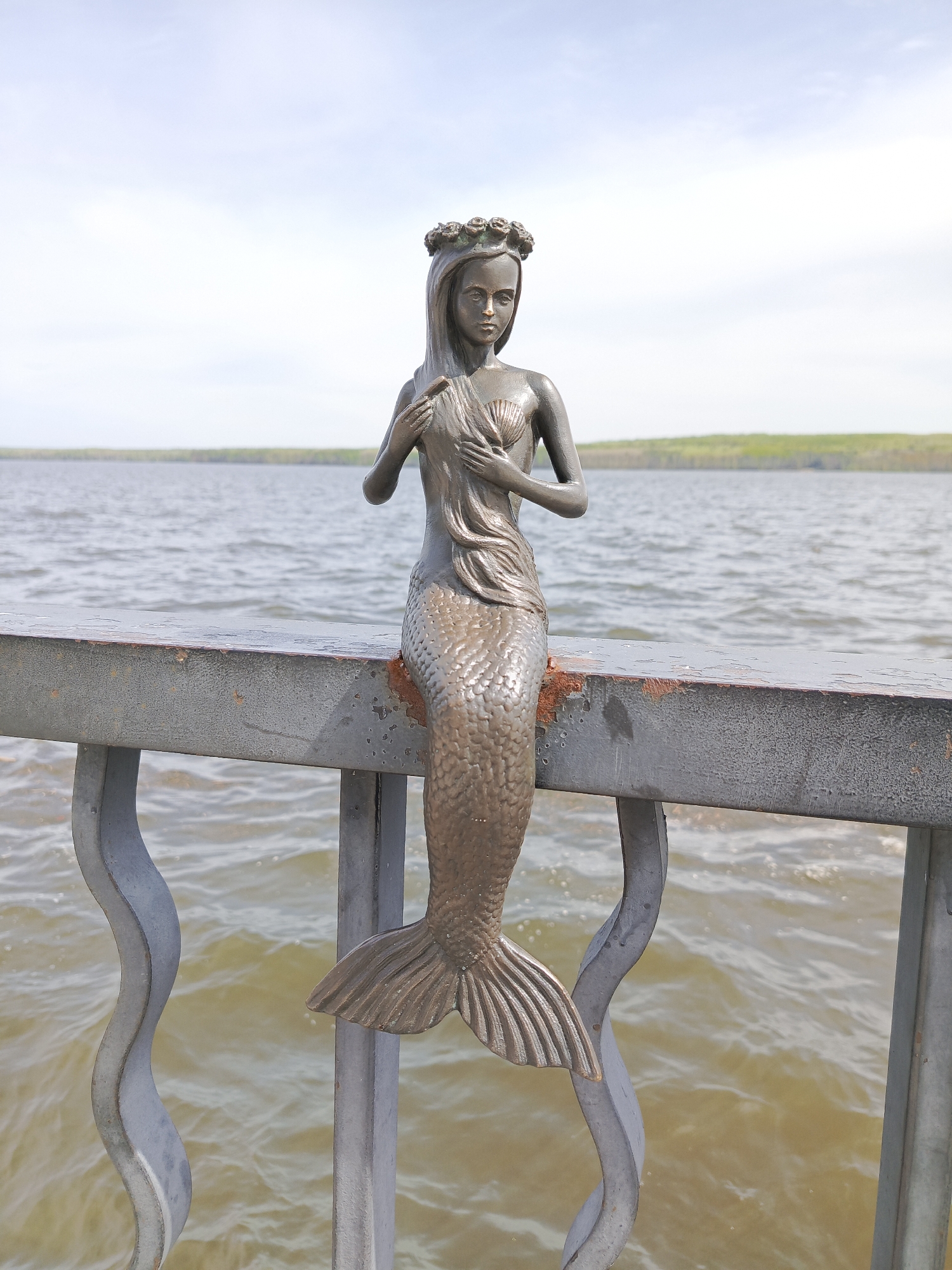 A bronze mermaid statue is perched on a gray, weathered metal railing. The statue depicts a young woman with long hair adorned with a floral crown, her hands gently placed over a seashell on her chest. Below her waist, she transforms into a scaly mermaid tail, the fin extending downwards between the bars of the railing. The railing is slightly rusted in places.
Behind the railing and the statue is a body of water, likely a lake or river, with gentle ripples on its surface. The opposite shore is visible in the distance, featuring a strip of green foliage beneath a light blue sky with wispy clouds.
The image likely depicts the mermaid statue located in the city of Chaikovsky, Russia.