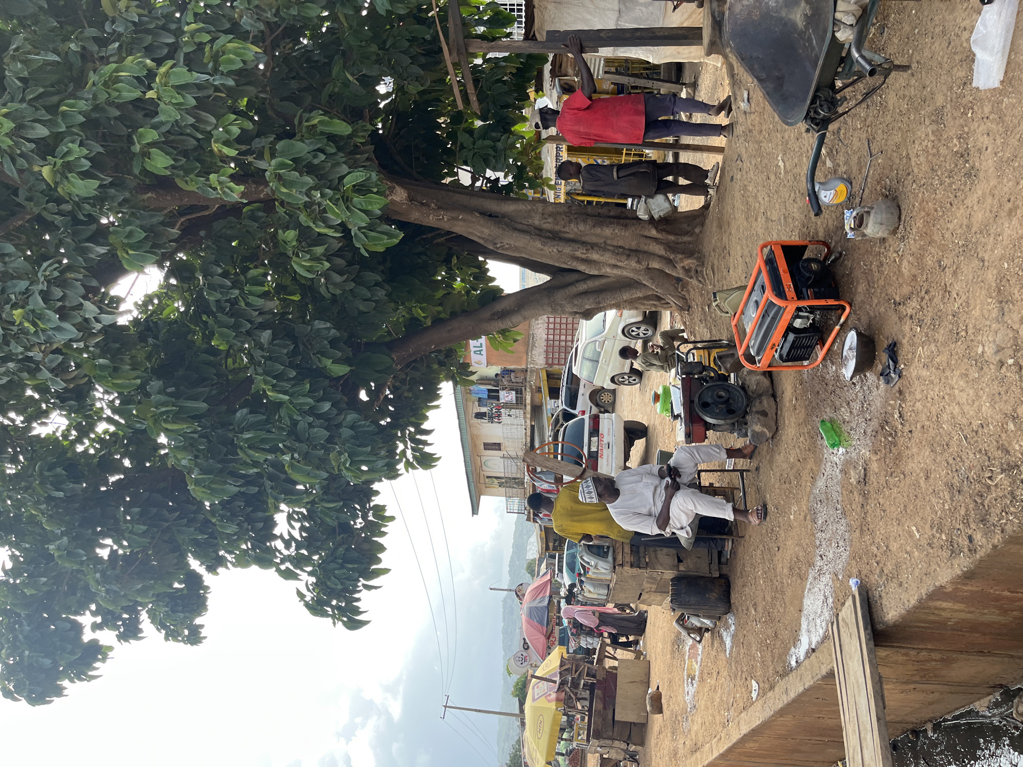 This image depicts a scene in Jos, Nigeria, likely a marketplace or workshop area. The focus is on a large, mature tree with dense green foliage, dominating the left side of the frame. Beneath the tree, several people are present. One man in a red shirt stands near the tree, holding onto a wooden pole. Another person in a dark jacket is also visible. Further to the right, an orange generator is a prominent object on the dusty ground, with an oil can and other tools nearby, suggesting mechanical work is being done or has recently occurred.
Several individuals are seated on makeshift benches or chairs to the right of the generator. One man, wearing a white traditional outfit and a cap, is seated and appears to be observing something. Another man in a yellow shirt sits next to him. In the background, vehicles, including white vans and cars, are parked. Buildings with signs and what appears to be clothing hanging to dry are visible behind the vehicles, indicating a commercial or residential area.
Further into the background, a series of stalls with umbrellas, some colorful, suggest a market setting. The terrain is predominantly dirt and sand. The sky is overcast with some bright patches, suggesting daytime with potentially mixed weather. The overall atmosphere seems to be one of casual daily activity. Visible text includes "ALL" on a building in the background, and partial text on a yellow sign which may be an advertisement. The scene appears to be outdoors, in a warm climate, possibly during the afternoon.