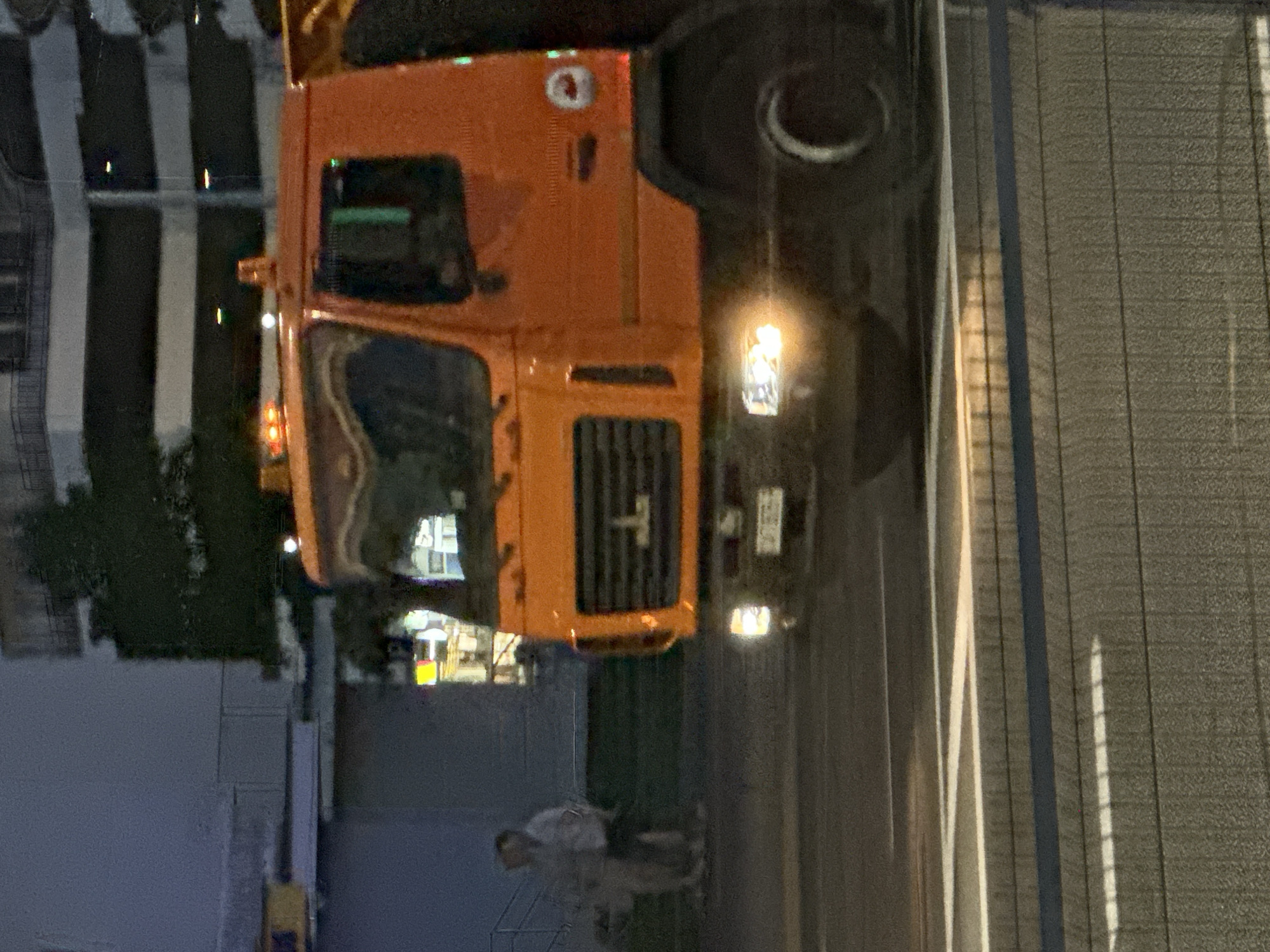 The image captures a close-up, side-view of an orange truck with its headlights on, in motion on a street at dusk or night. The truck's front grille, windshield, and right side are visible. The vehicle is positioned in the foreground, with its front tire and part of the undercarriage also in view. In the background, two indistinct figures, appearing as a man and a woman, are standing on what looks like a sidewalk or grassy area. Behind them, a building with multiple floors and a tree are partially visible. The environment suggests an urban setting with streetlights or illuminated signs contributing to the ambient light. The street is paved, and a sidewalk or curb is present between the roadway and the background elements.