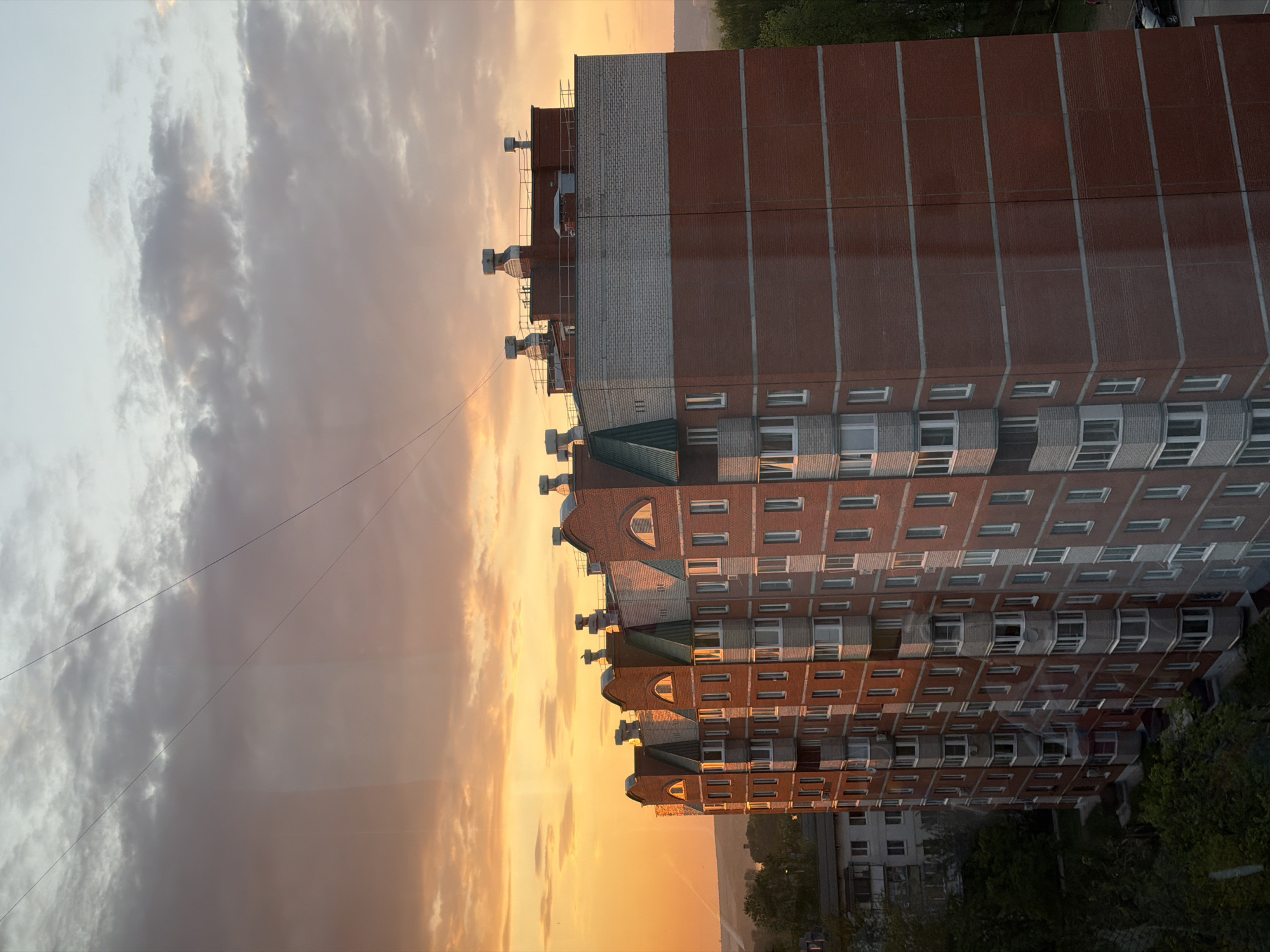 The image is an upward-angle view of an apartment building in Chaikovsky, Russia, at sunset. The building is brick with white trim around the windows and accents. The architecture includes arched details on the upper floors. To the left of the building is a sky filled with orange and gray clouds indicative of a sunset. Power lines are visible crossing the sky. Green trees are visible at the bottom and top edges of the frame.