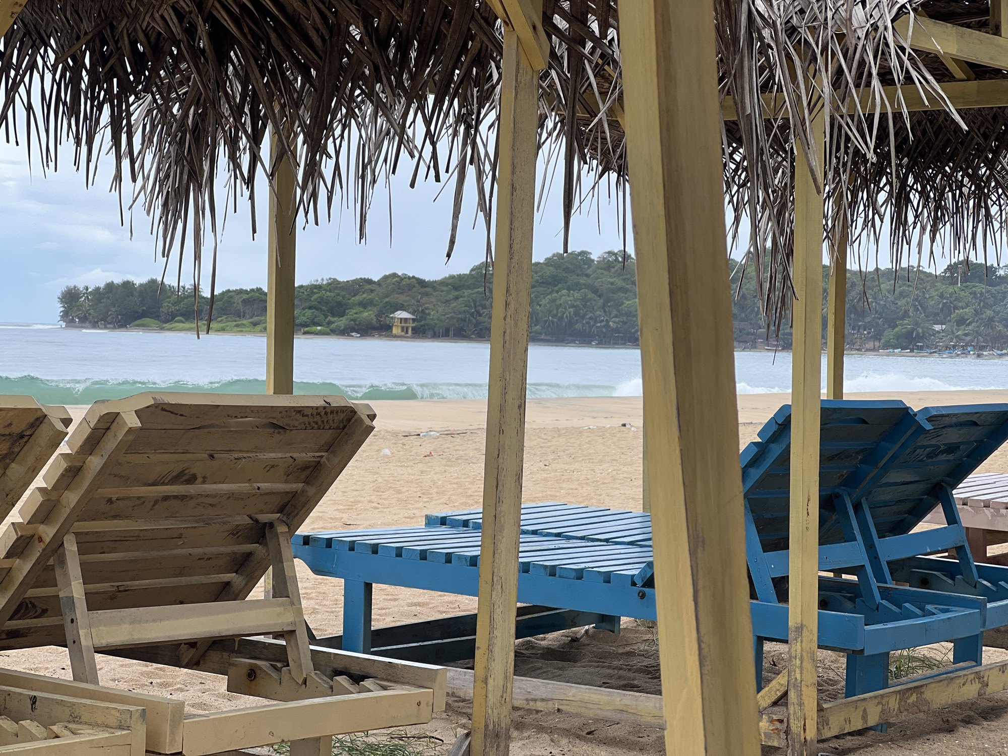 This image captures a serene beach scene viewed from beneath a thatched-roof structure, likely a cabana or beach shelter, in Arugam Bay, Sri Lanka. The foreground is dominated by the rustic texture of dried palm fronds forming the roof, which hangs down, partially obscuring the view and creating a natural frame. Light yellow, weathered wooden support beams for the structure are prominent, some vertically aligned in the center and right.
On the sandy ground within the shelter, several wooden beach loungers are visible. To the left, one unpainted, natural wood-colored lounger is seen from the back, its headrest in an upright position. To the right, two blue-painted wooden loungers are visible, also angled upwards, partially behind a wooden pillar.
Beyond the immediate shelter and loungers, the sandy beach stretches out to the ocean. Gentle waves can be seen rolling in and breaking near the shore. The sky is overcast, suggesting a cloudy day without direct sunlight. In the distance, a lush green coastline with dense tropical vegetation curves around the bay. A small, distinctive light yellow building with a prominent roof stands out on the distant shore amidst the trees.
The scene is devoid of people or discernible activity, conveying a peaceful and quiet atmosphere. The tropical foliage, thatched roof, and beach setting are strong indicators of a resort or coastal area. No visible text is present in the image.