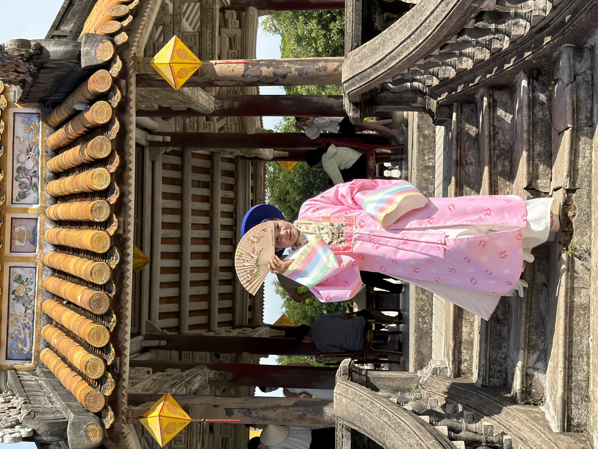 A person wearing a pink traditional Vietnamese ao dai and a blue hat stands on a stone staircase, holding an open fan. The person is positioned centrally in the frame, with the staircase leading up to a structure with a tiled, yellow, pagoda-style roof. Yellow lanterns are suspended from the roof. The background shows other people and trees. The scene appears to be in a historic or cultural site, potentially within Phú Xuân Ward, Vietnam, given the architectural style.