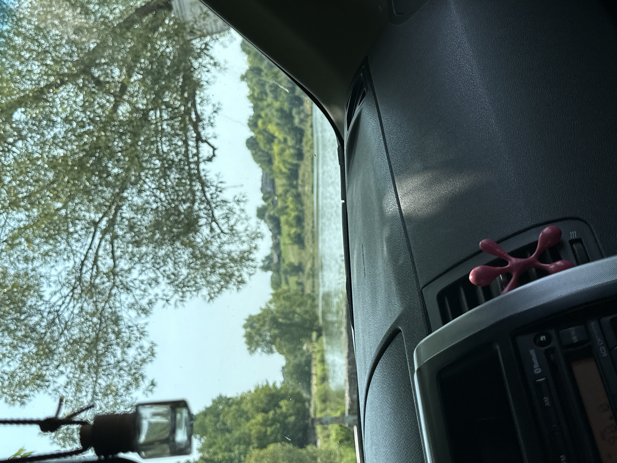 The image is taken from inside a car, looking out through the windshield. In the foreground, the dashboard of the car is visible, with a portion of the center console and air vent. A small, pink, octopus-shaped air freshener is attached to the air vent. Hanging from the rearview mirror is a small glass bottle, likely an air freshener as well.
Outside the car, a calm body of water, possibly a lake or pond, is visible. Trees and green hills form the background, with some houses sparsely scattered amongst them. The sky is bright and clear, indicating daytime. The location is identified as Makiv, Ukraine.