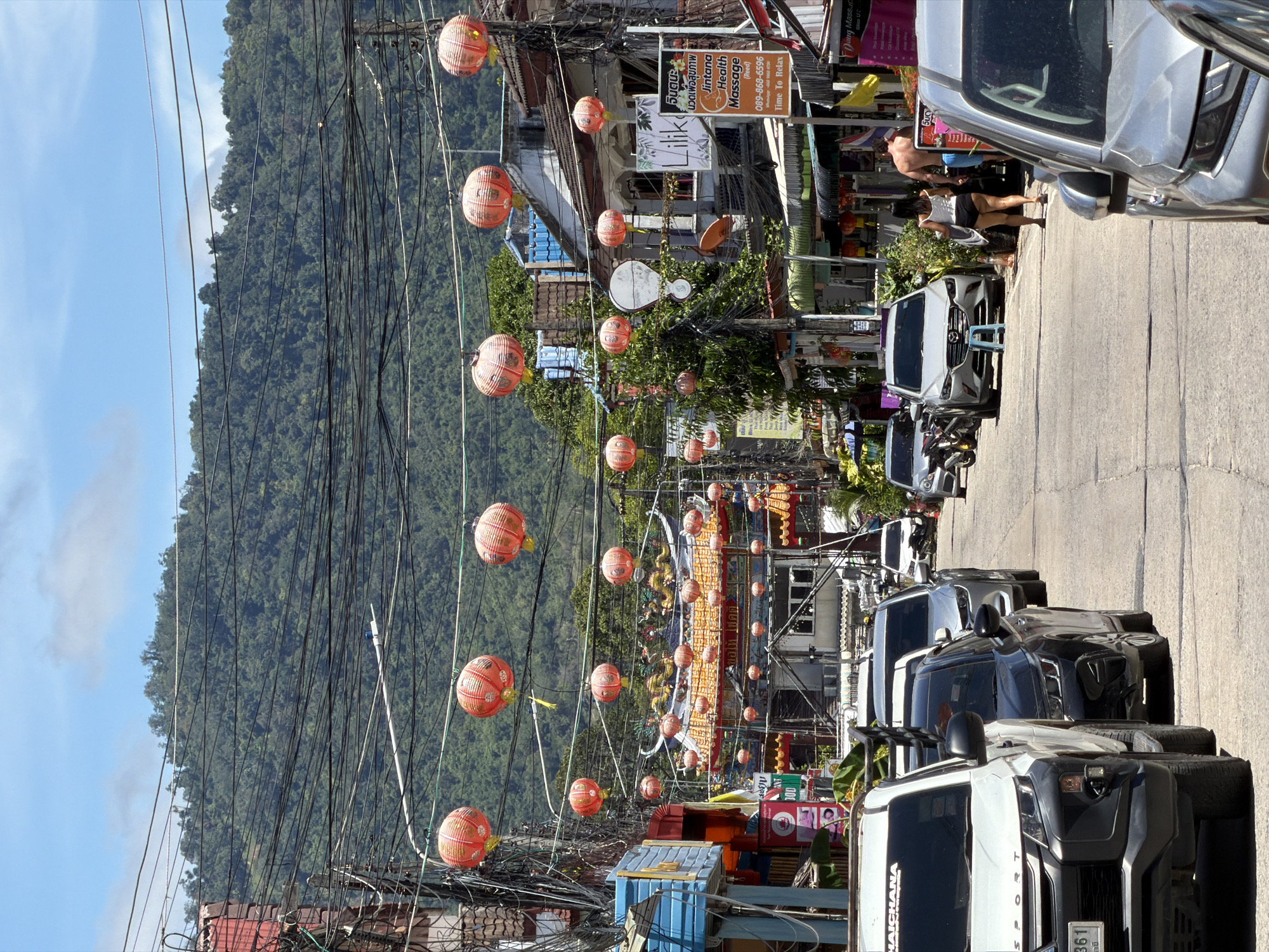 The image depicts a street scene in Baan Mae Nam, Thailand, characterized by a dense network of overhead wires. Red Chinese lanterns are suspended across the street, strung between buildings and poles, with some appearing to be lit. Buildings lining the street exhibit various architectural styles and colors, with some featuring awnings and signs in Thai script. A prominent building with a traditional Chinese-style roof, adorned with dragon motifs, is visible in the mid-ground. Cars are parked and driving on the paved road, including a white pickup truck in the foreground. In the background, a lush green hillside covered in trees rises steeply. Two individuals, one wearing a white top and dark shorts and the other shirtless in blue shorts, are walking away from the camera on the right side of the street. The sky is blue with scattered white clouds. The overall atmosphere suggests a bustling commercial area with a strong cultural influence.