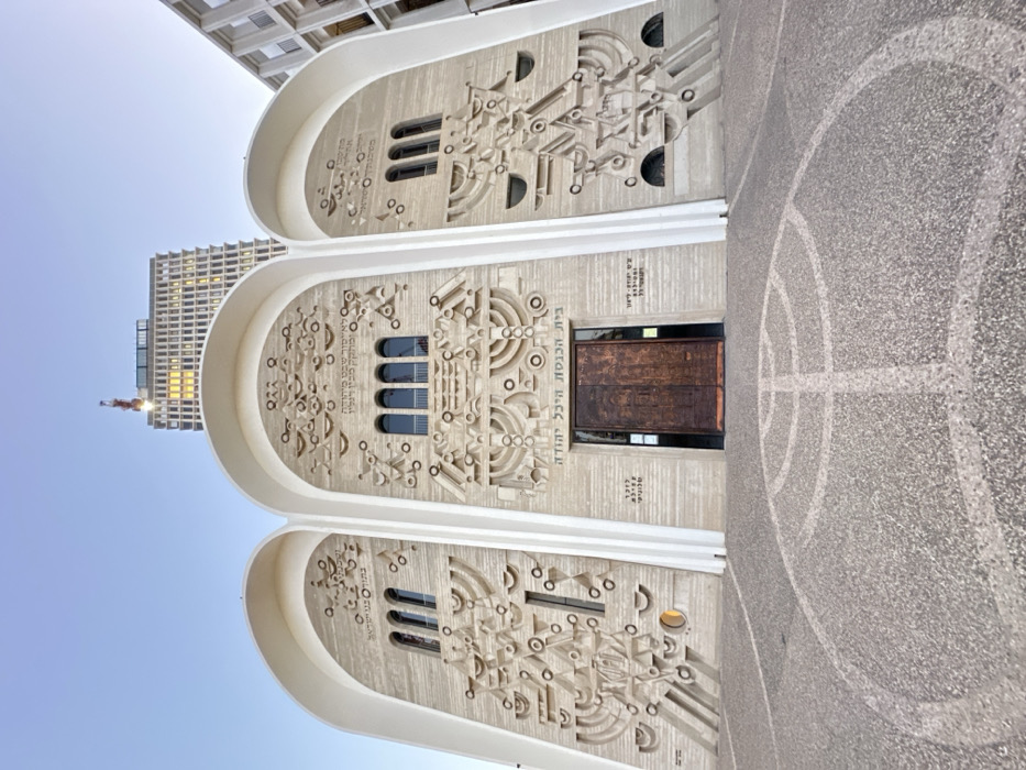 The image depicts the Heichal Yehuda Synagogue, also known as the Kalmania Synagogue, located on Ben Yehuda Street in Tel-Aviv, Israel. The synagogue's facade features three prominent, light-colored arched sections over a textured stone or concrete surface. Elaborate bas-relief sculptures adorn the walls within these arches, incorporating stylized menorahs, Stars of David, and geometric patterns. Multiple dark-paned windows are integrated into these relief designs.
A large, double-leaf, dark-toned metal or wood door serves as the main entrance, centrally positioned beneath the middle arch. Above this door, the Hebrew inscription "בית הכנסת היכל יהודה" (Synagogue Heichal Yehuda) is visible, with additional Hebrew text inscribed on other sections of the facade.
The ground directly in front of the synagogue entrance is paved with light-colored aggregate, featuring a large circular pattern inlaid into the surface, which includes a stylized menorah design.
In the background, a tall, modern high-rise building with a grid of windows, some reflecting light, rises directly behind the synagogue. A red and white construction crane is visible atop this high-rise. To the right of the synagogue, part of another modern building with repetitive architectural elements is partially visible. The sky overhead is clear and light blue. No individuals are present in the scene.