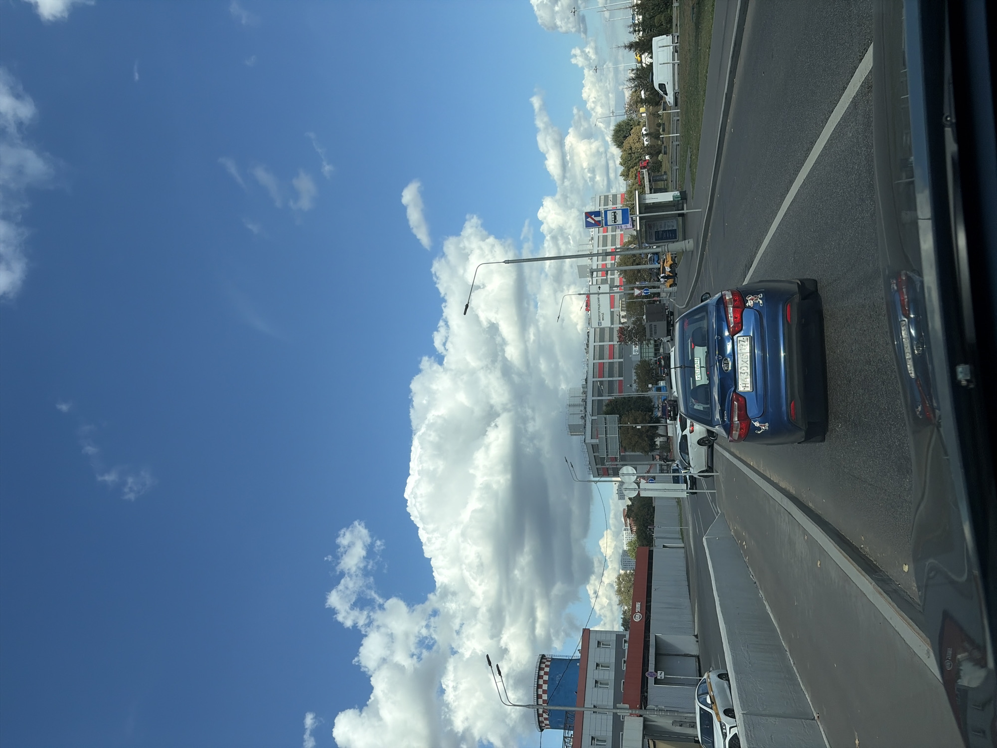 The image shows a street scene in Moscow, Russia, on a sunny day with scattered clouds. The perspective is from inside a vehicle, looking forward. In the foreground, the hood of the car is visible. In the middle ground, several cars are on the road, including a blue Kia sedan in the center lane. The car has a sticker on its rear bumper. To the left, a white car with a yellow stripe is parked. Further down the road, there is a bus stop with a blue sign. On the right side of the road, a white van is visible. In the background, there are buildings, including a large, modern shopping center with red and white accents. There are also streetlights and trees lining the road. The sky is a clear blue with large, fluffy white clouds. The scene appears to be a typical urban environment with ongoing traffic.