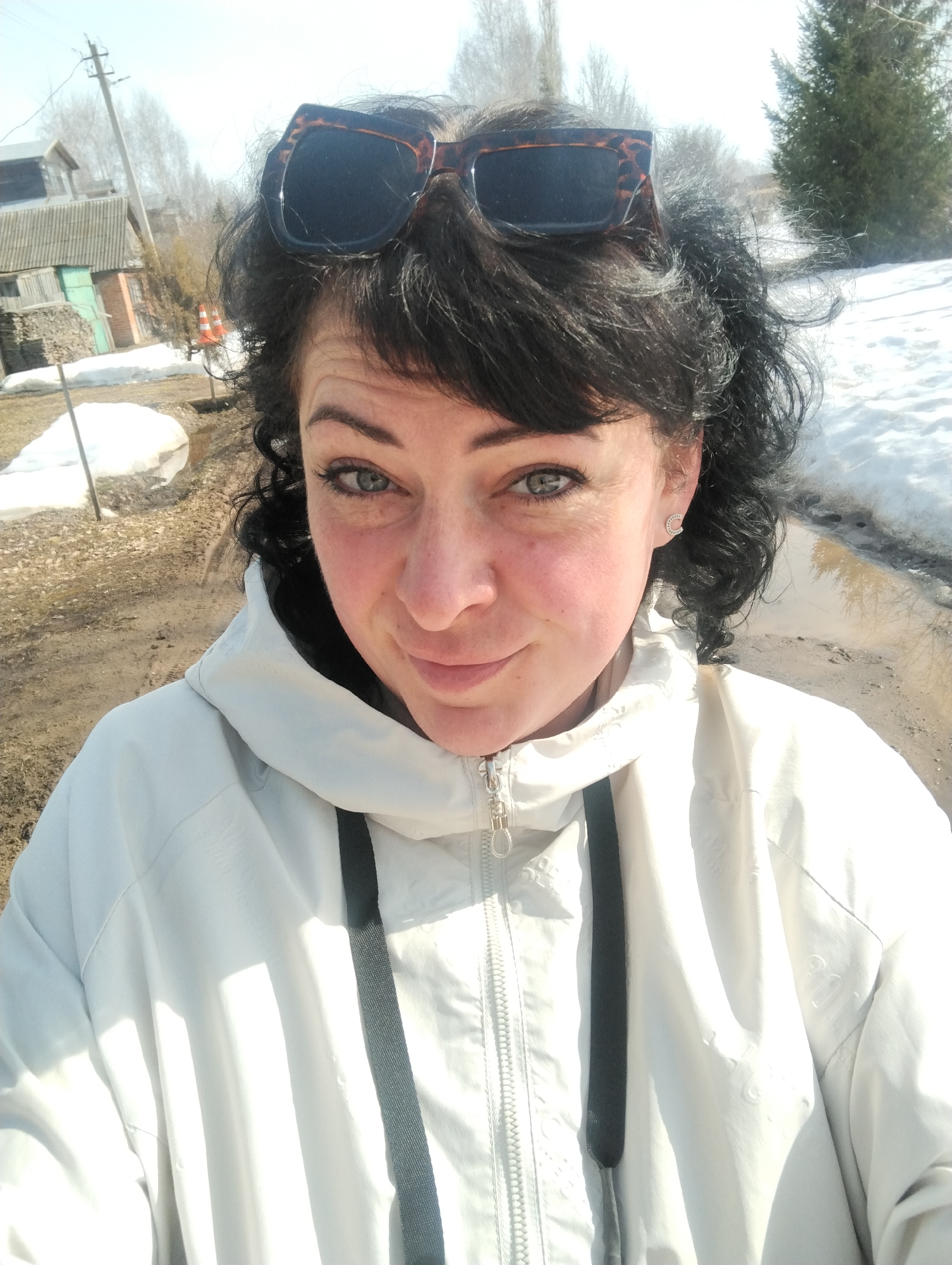 A woman with dark curly hair and sunglasses perched atop her head is centered in the frame. She is wearing a light-colored hooded jacket with a zipper and dark straps. Her face is flushed, and she looks directly at the camera.
Behind the woman, the scene is an outdoor environment. There is melting snow on the ground, and a muddy path or road is visible. To the left, there is a house with a green-painted section and stacked firewood, and further back, power lines and bare trees. To the right, there is a large evergreen tree.
The scene's location is in the city of Grishanki, Russia. The picture appears to be taken on a sunny day.